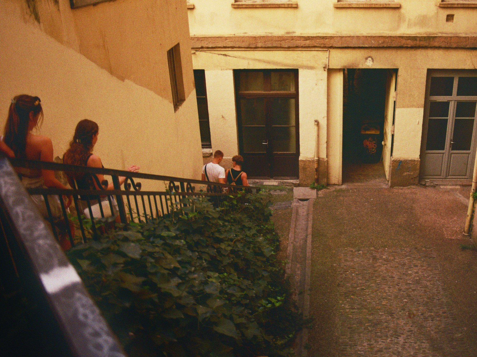 Vier Menschen sitzen auf einer Balkonbrüstung, während zwei weitere Personen durch eine Tür im Hintergrund in einem Innenhof gehen.