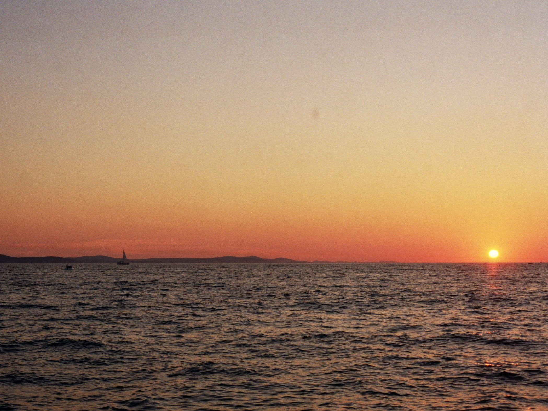 Sonnenuntergang über dem Meer mit Segelbooten und entfernten Hügeln im Hintergrund
