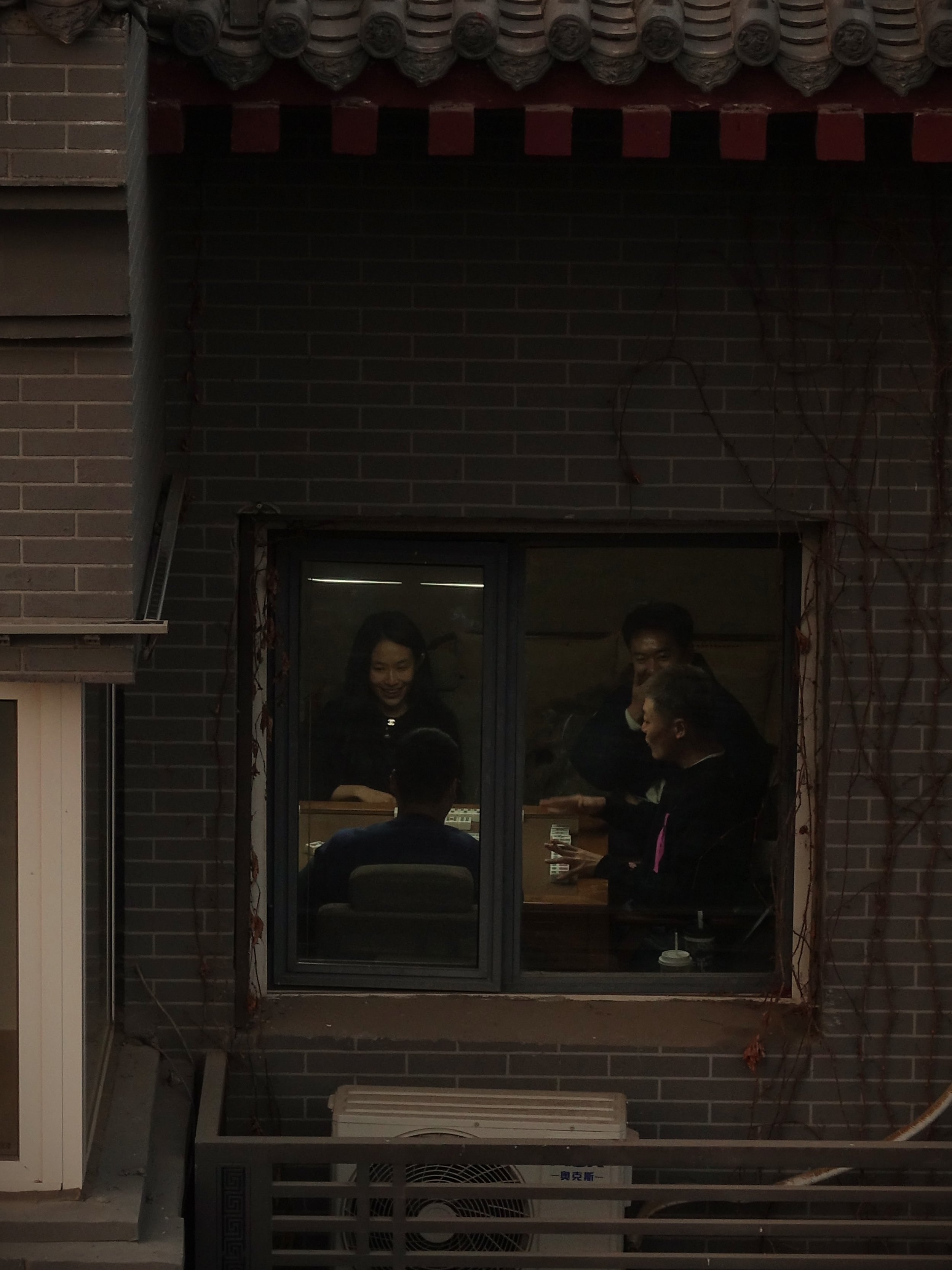 Menschen sitzen und stehen an einem Tisch im Inneren eines Raums, sichtbar durch ein Fenster in einem Brickwall-Gebäude, während sie sich unterhalten.