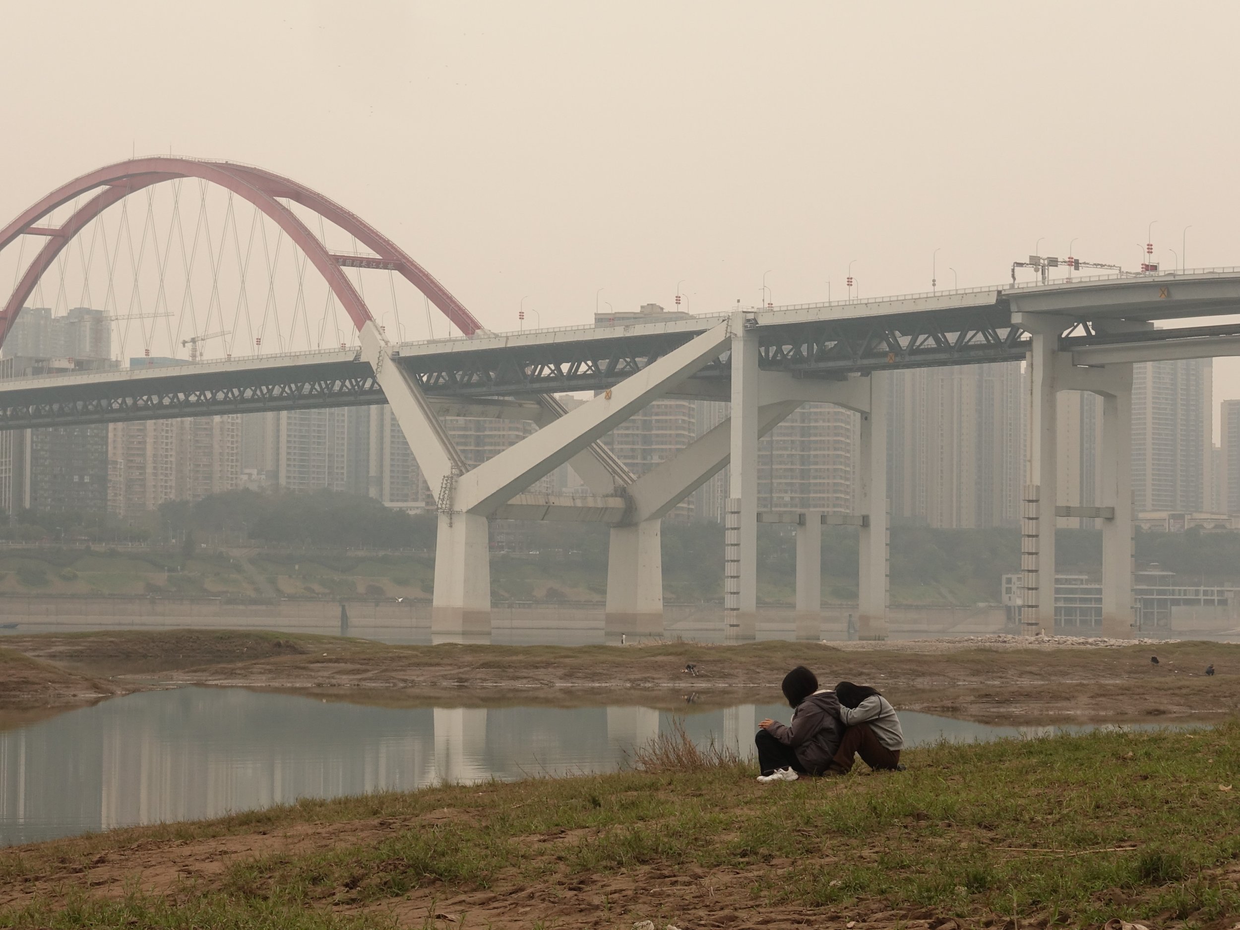 Zwei Kinder sitzen am Ufer eines Flusses, im Hintergrund eine große Brücke mit Hochhäusern in der Stadt. Die Kinder scheinen entspannt und blicken auf den Fluss.