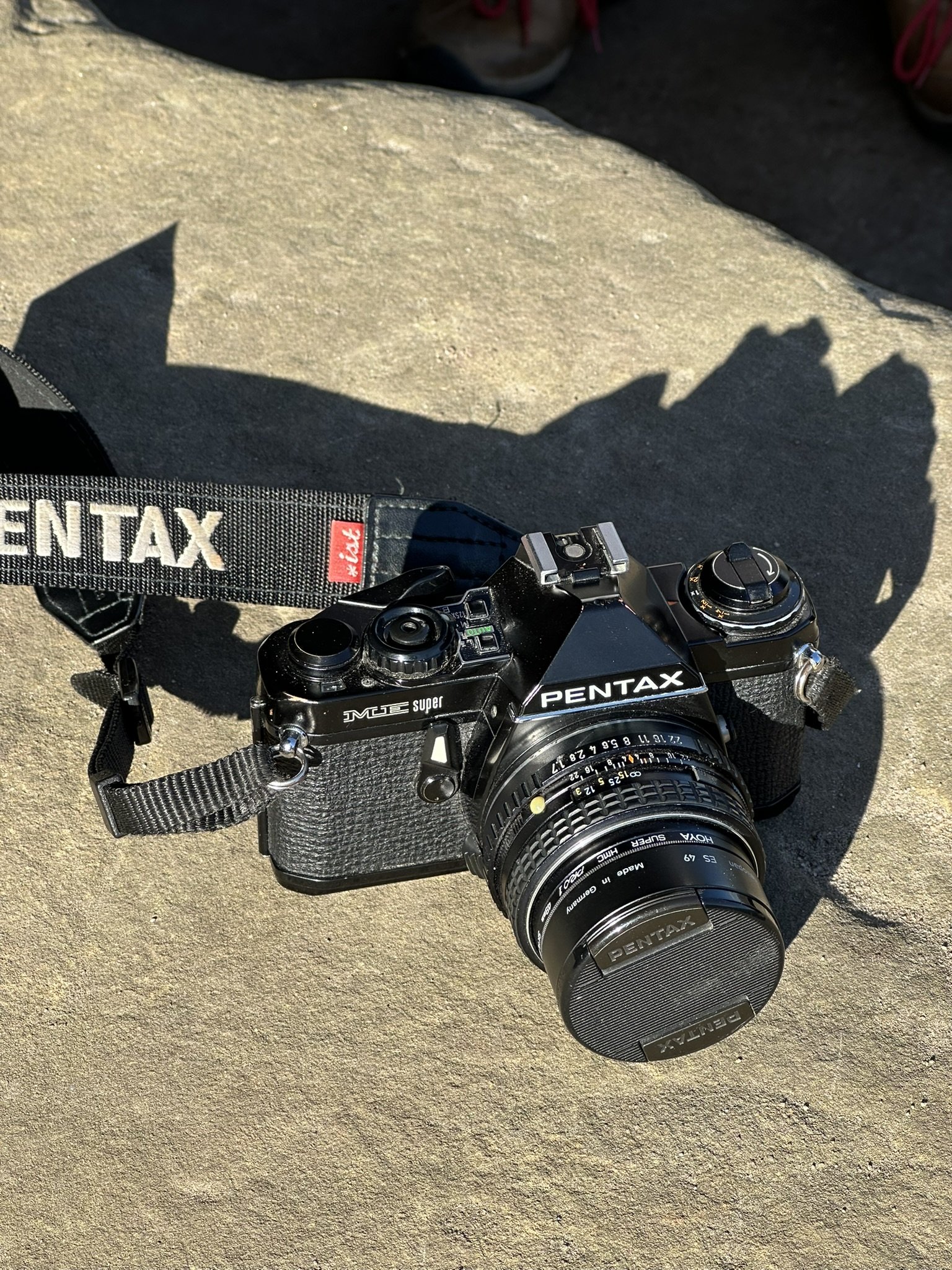 Eine Kamera der Marke Pentax liegt auf einem grauen Stein im Sonnenlicht.