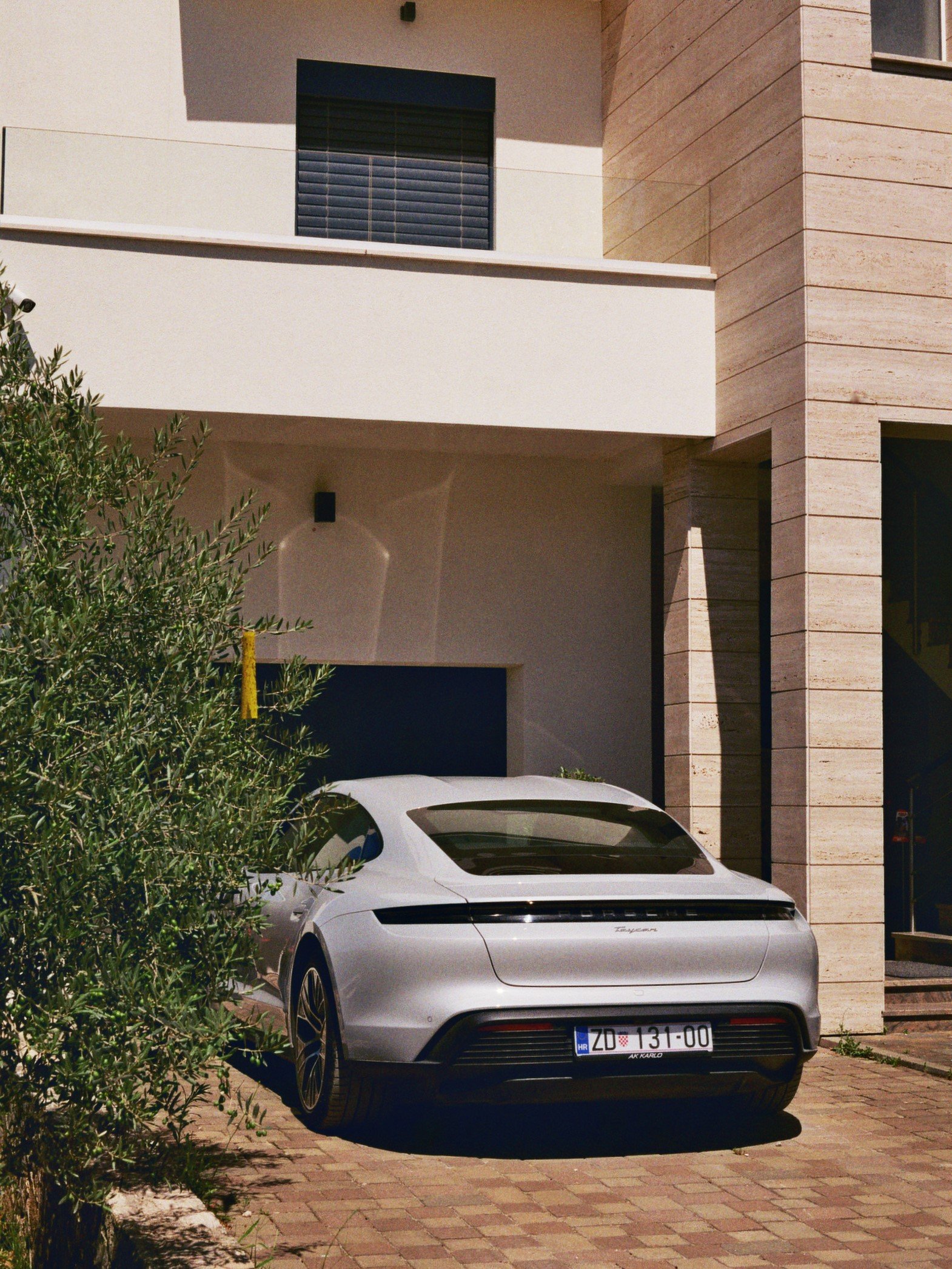Ein silbernes Auto, vermutlich ein Porsche Taycan, parkt vor einem modernen, mehrstöckigen Wohnhaus mit weißen und beige Farben. Es ist sonnig, und das Auto steht auf einem gepflasterten Bereich.