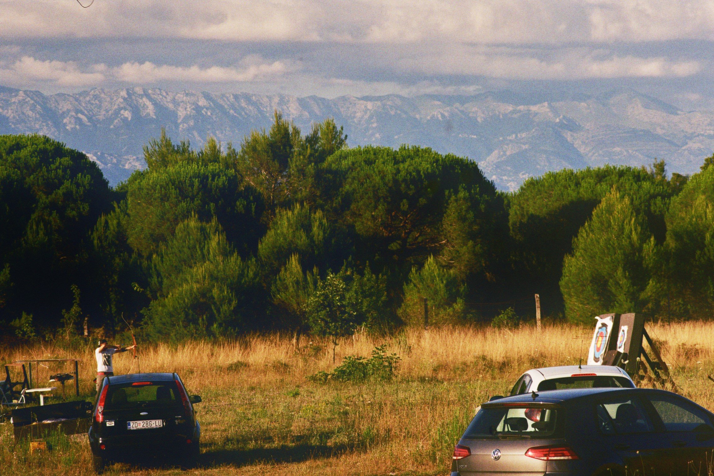 Junge beim Bogenschießen auf einer Wiese mit mehreren Autos und Zielscheiben, im Hintergrund Bäume und Bergketten.