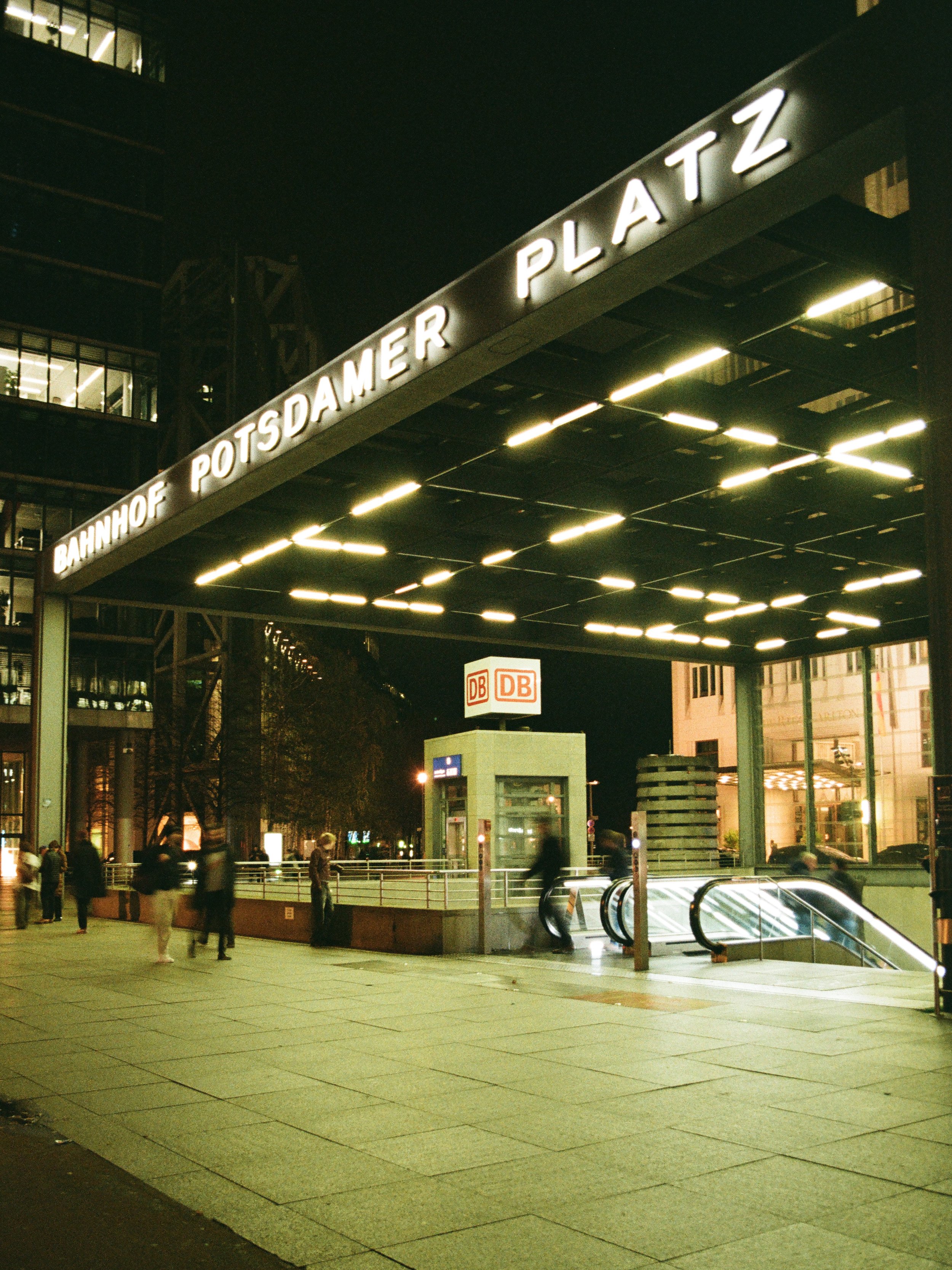 Am Abend beleuchteter Eingang zum Potsdamer Platz Bahnhof in Berlin, mit Menschen, die die Treppe hinauf- und hinuntergehen, und den Schildern der Deutschen Bahn.