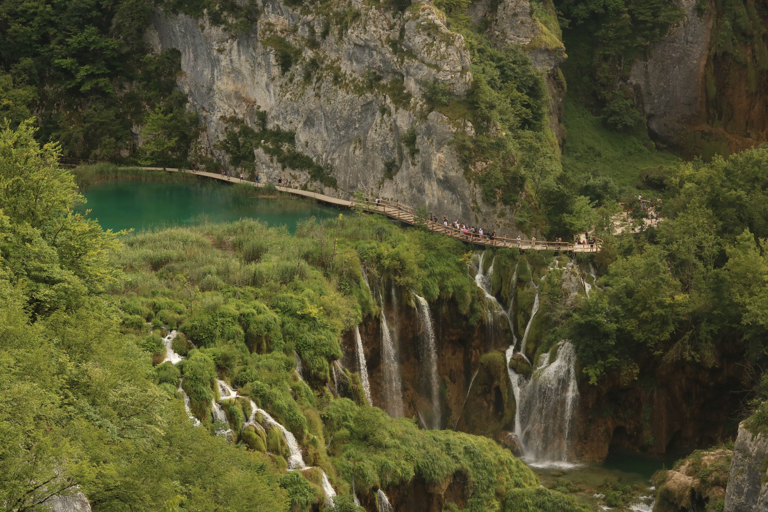 Ein grüner Wasserfall mit mehreren kleinen Wasserfällen, umgeben von üppigem Grün und steilen Felswänden. Es gibt einen Weg, auf dem Menschen spazieren, der entlang des Wasserfalls führt.