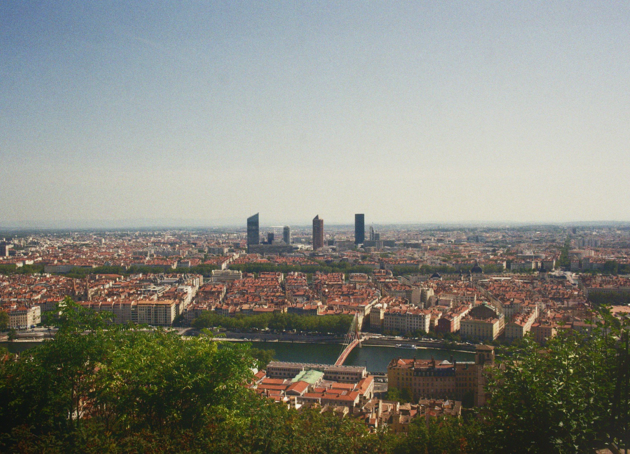 Panorama von Paris mit Fluss, Brücken, Gebäuden und Hochhäuser im Hintergrund, grüne Bäume im Vordergrund bei Tageslicht.