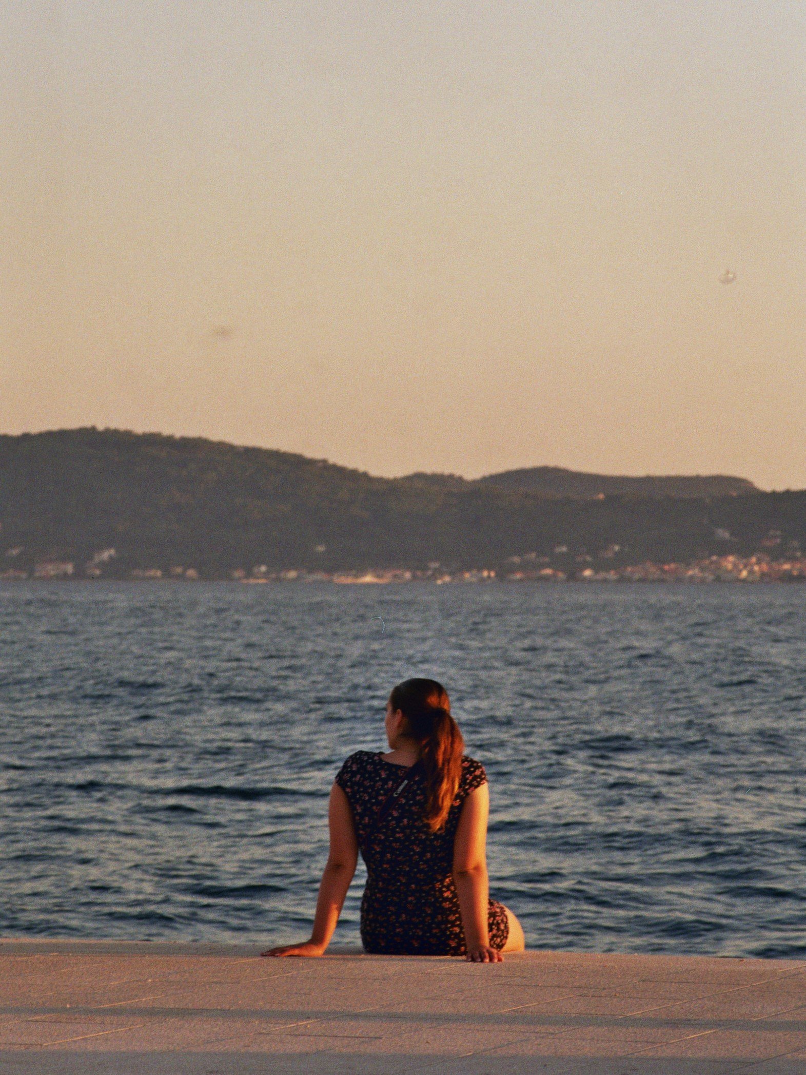 Junge Frau sitzt auf einer Uferpromenade mit Blick auf das Wasser und Berge im Hintergrund, Sonnenuntergangsstimmung.
