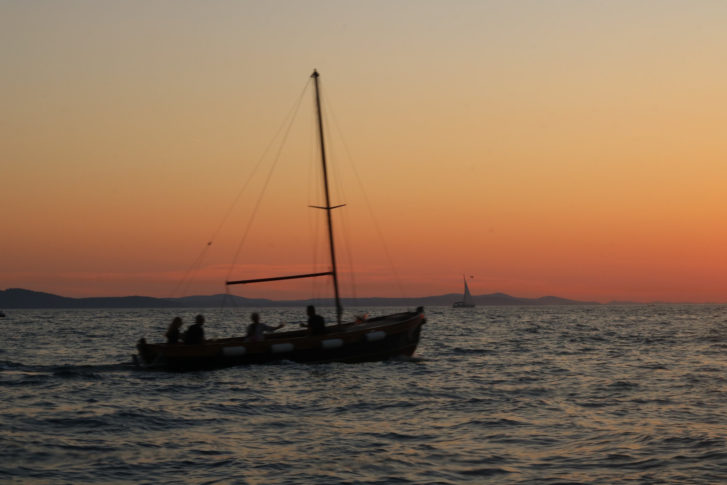 Segelboot mit Passagieren auf dem Wasser bei Sonnenuntergang, im Hintergrund entfernte Segeljachten und Inselkonturen.