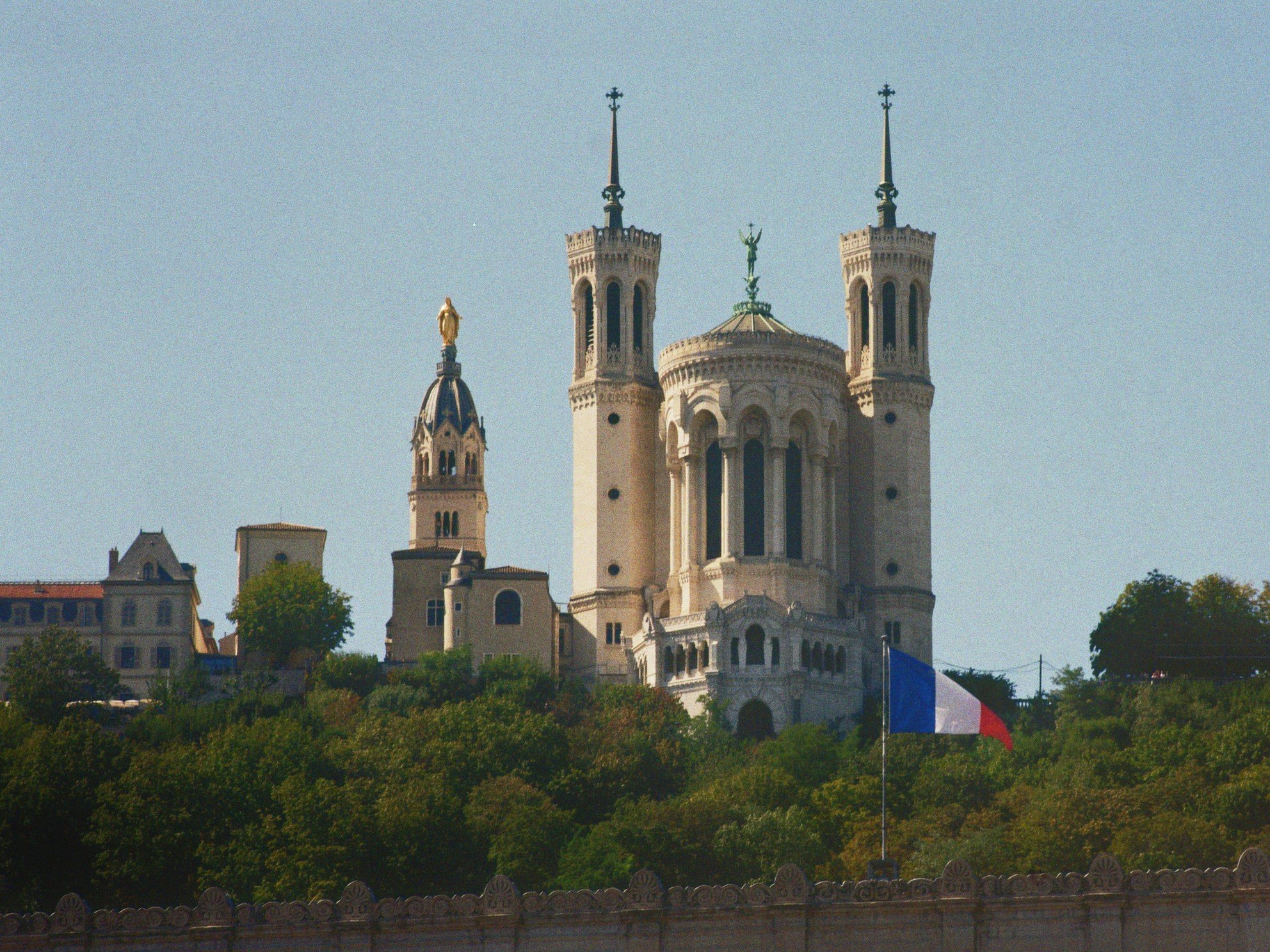 Christusstatue auf einer Kirche mit zwei Türmen, französische Flagge im Vordergrund, umgeben von Bäumen.