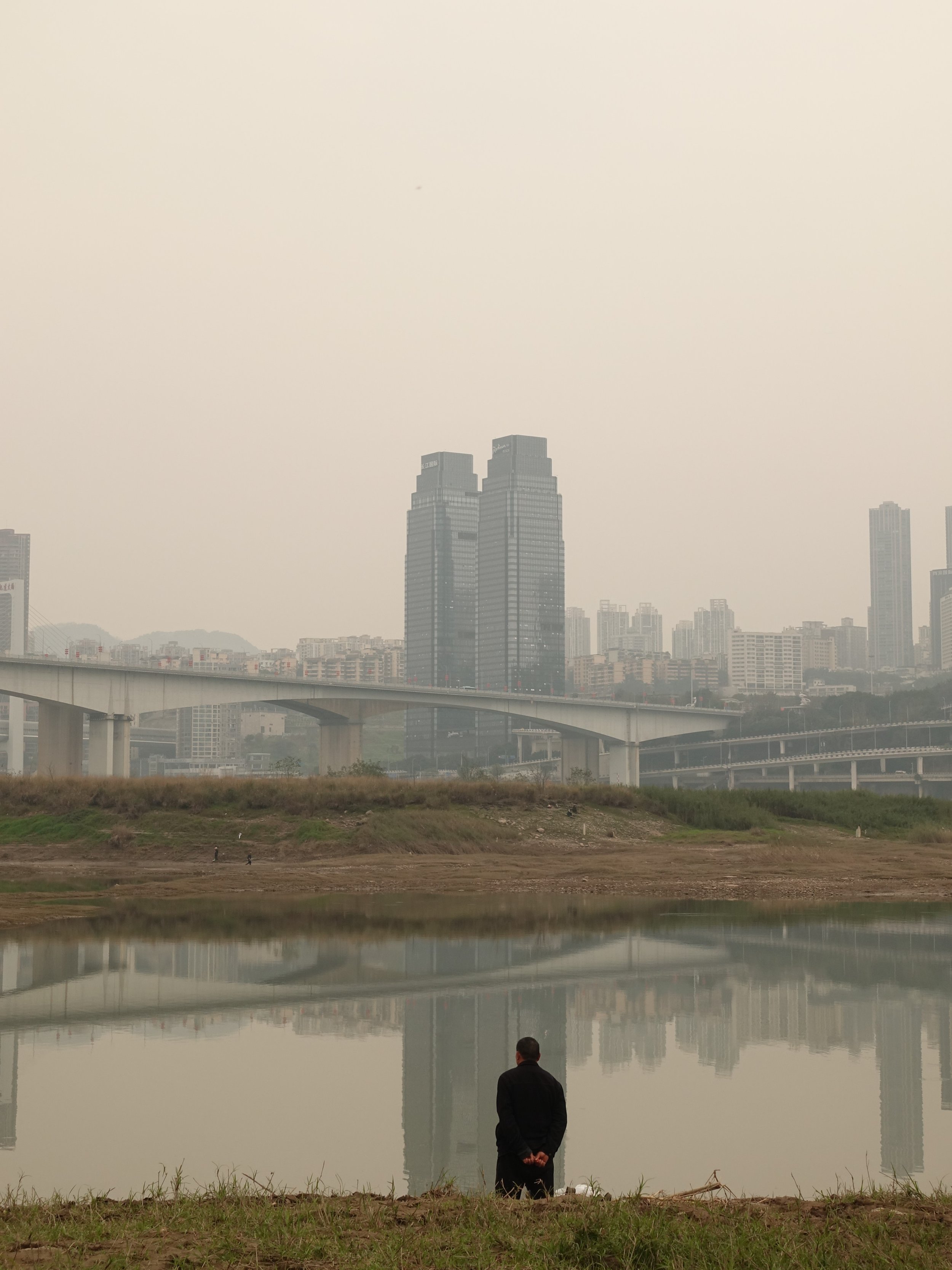 Ein Mann steht am Ufer eines Flusses oder Sees, blickt auf die Wasserfläche, mit einer Stadt im Hintergrund, bestehend aus hohen Gebäuden und Brücken, bei bewölktem Himmel.
