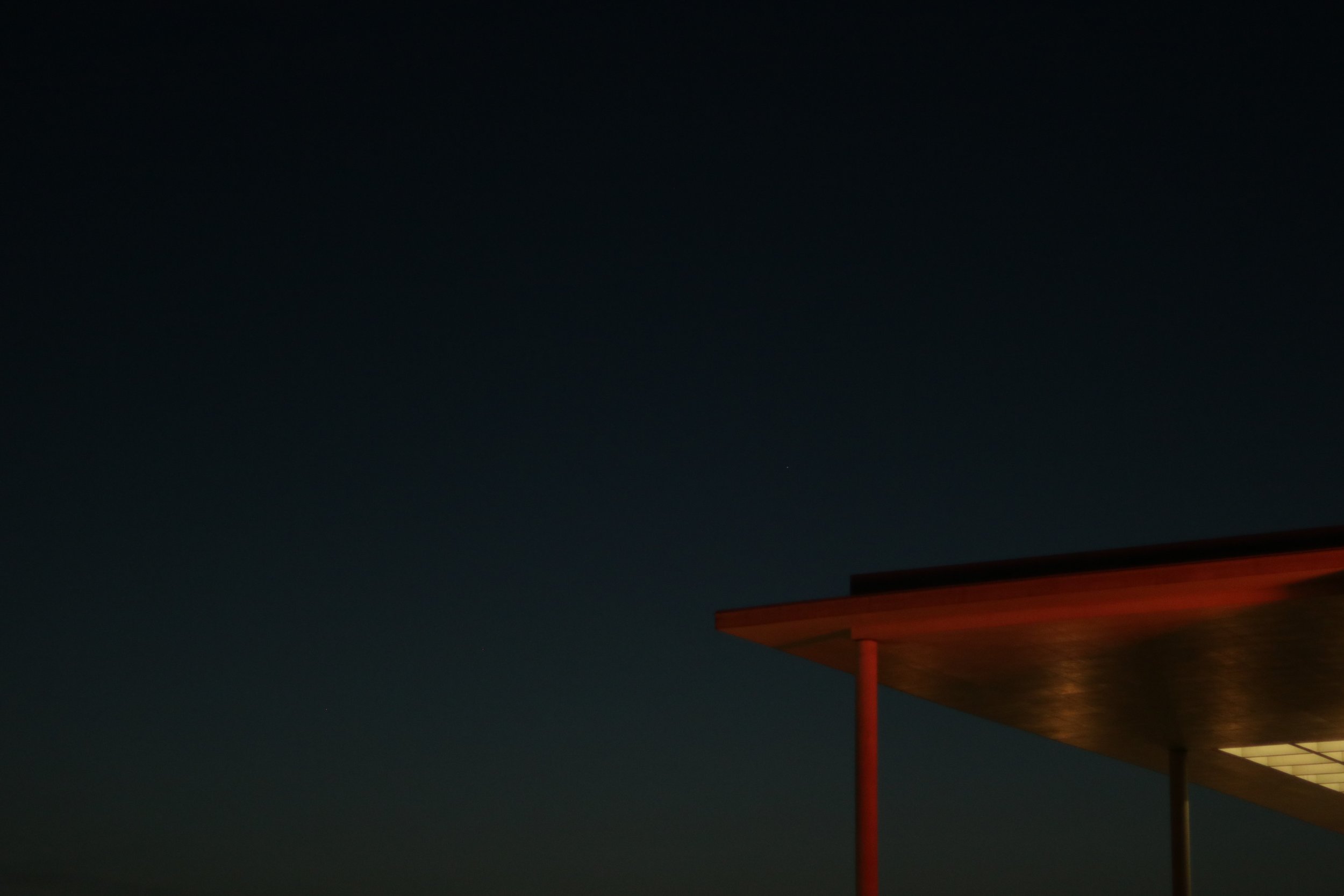 Nachtaufnahme eines modernen Gebäudes mit der rötlich beleuchteten Dachkante, sichtbarer Unterstützungssäule, gegen dunklen Himmel.