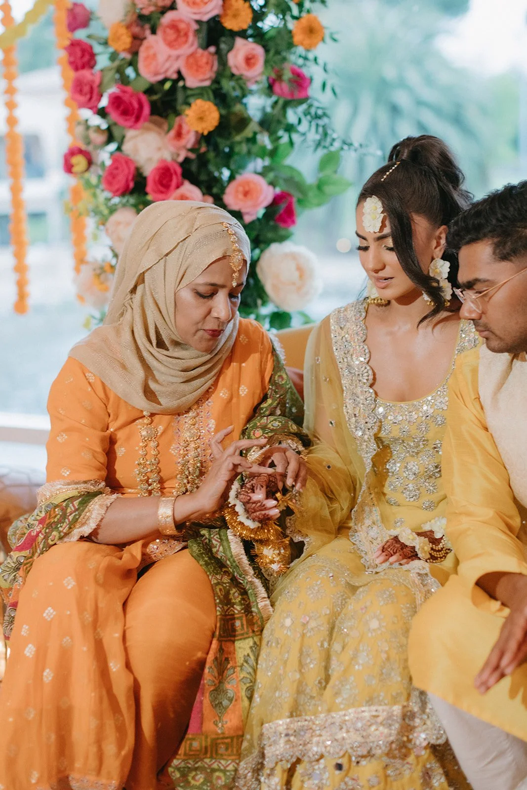 Three people dressed in traditional Indian clothing, seated together during a celebration with a floral backdrop.
