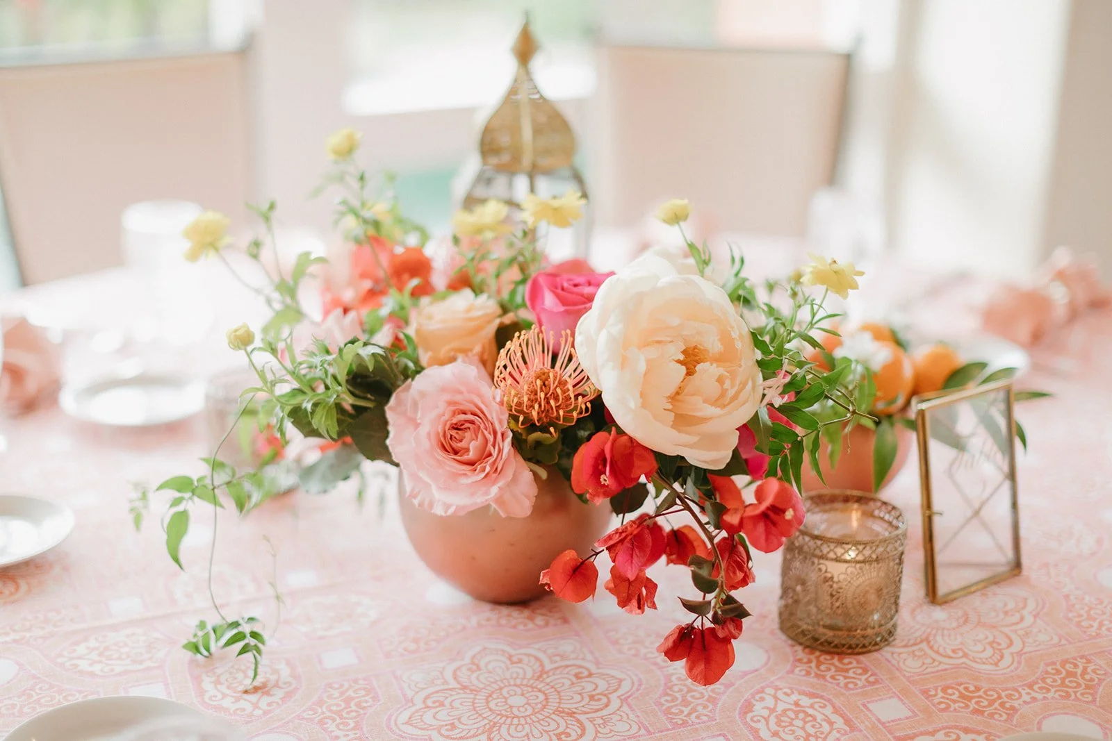 Elegant table centerpiece with pink, white, and peach flowers in a round vase, surrounded by gold candle holders and glassware on a patterned pink tablecloth.
