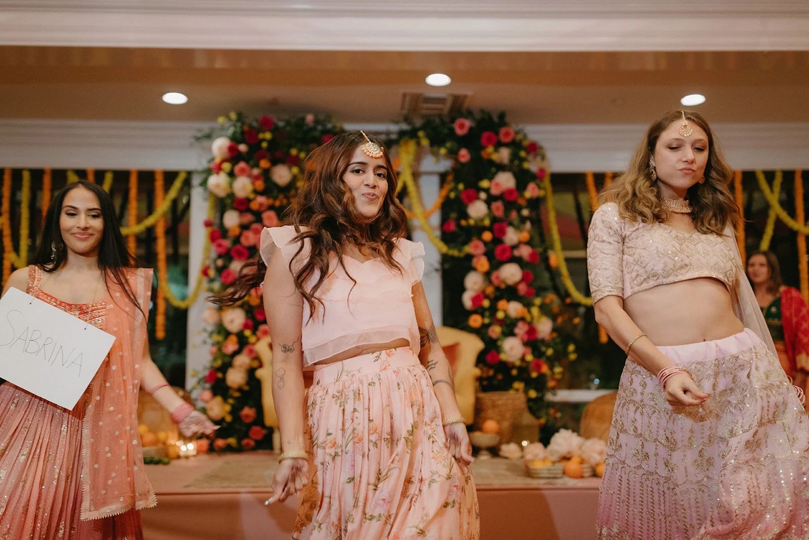 Women dressed in traditional Indian clothing performing a dance at a decorated event with flowers.