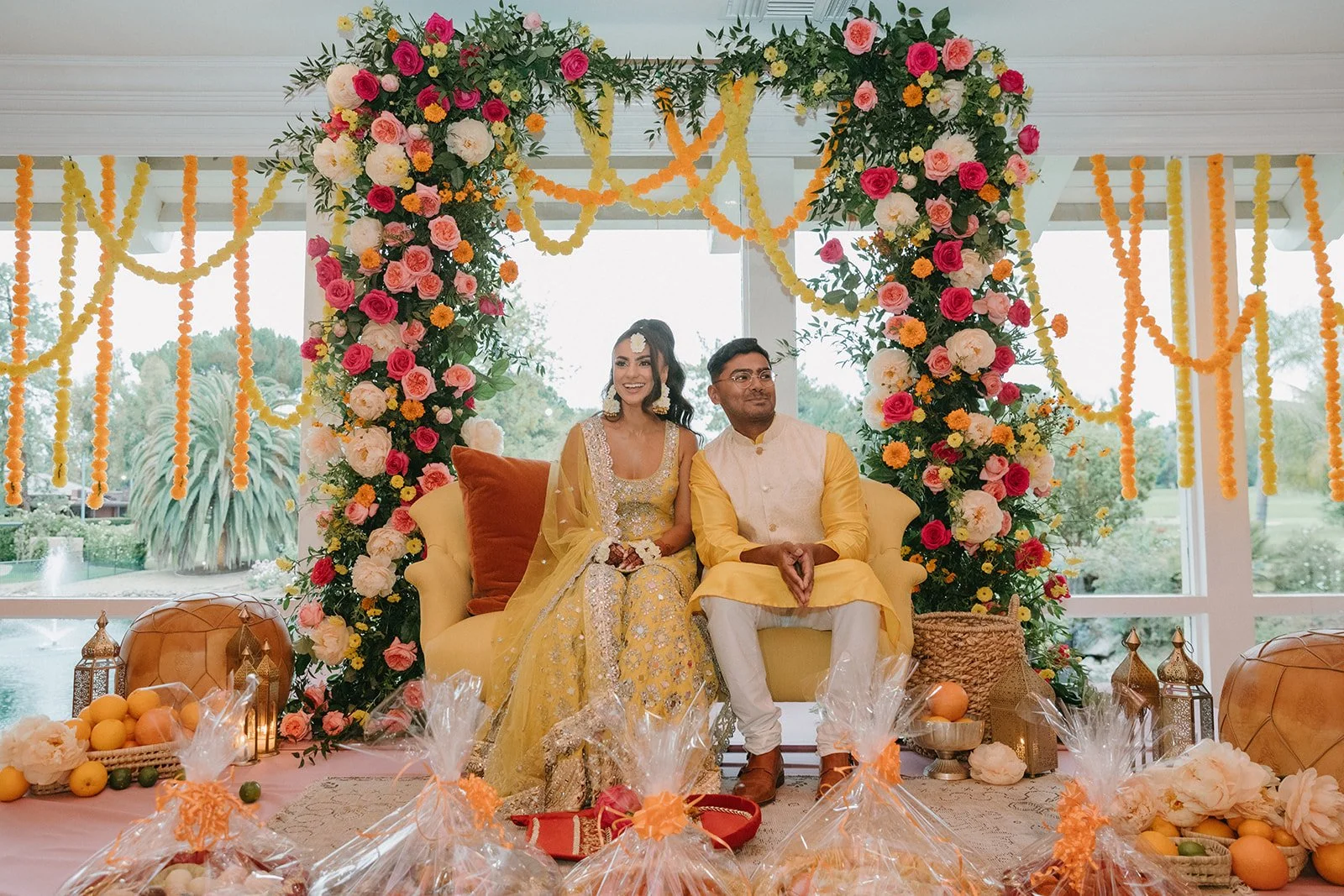 A couple dressed in traditional Indian attire sitting on a yellow sofa at a wedding or engagement ceremony, surrounded by a floral arch with pink, white, and yellow flowers, and decorative elements like fruits and gifts in the foreground.