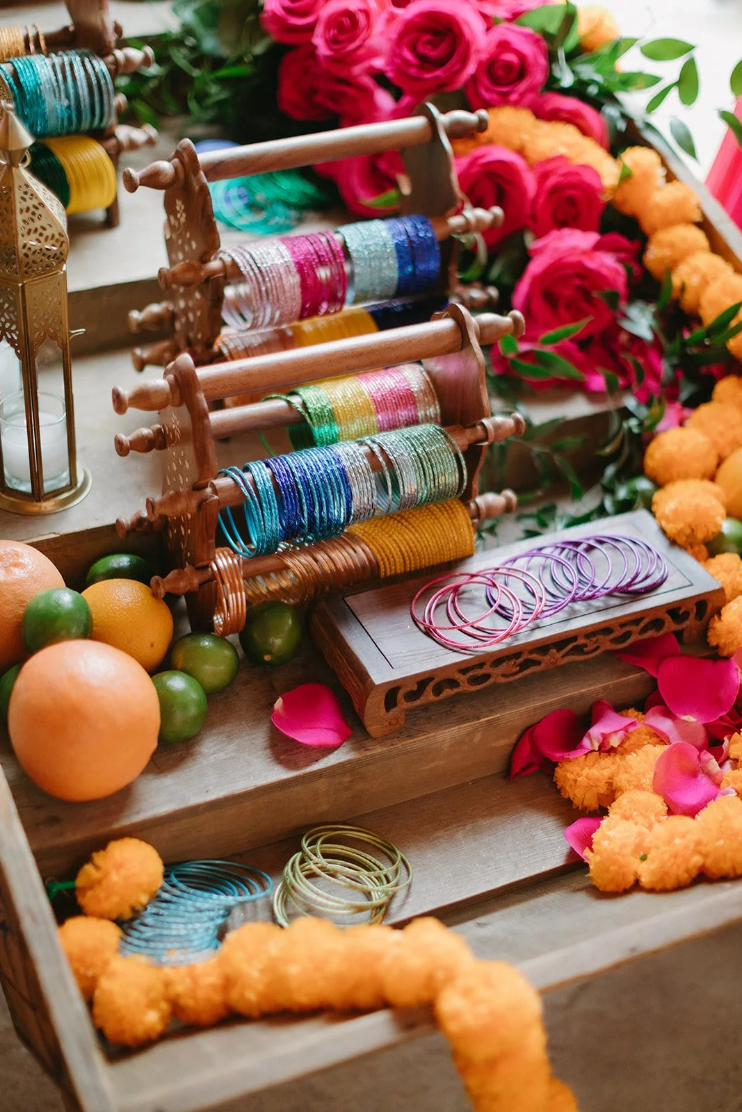 Colorful bangles and bracelets on wooden display stands, surrounded by oranges, limes, pink roses, marigold flowers, and pink petals.