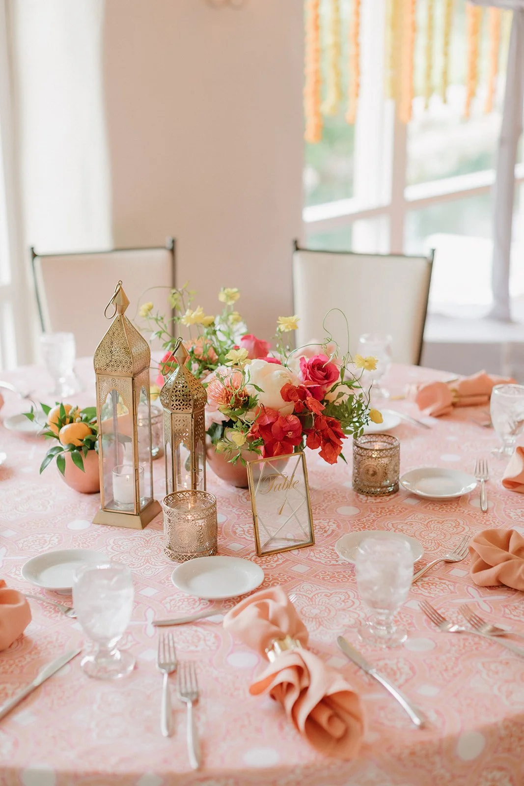 Elegant round table decorated with pink and floral centerpieces, candles, table number, and set with plates, silverware, napkins, and glasses in a bright room with windows.