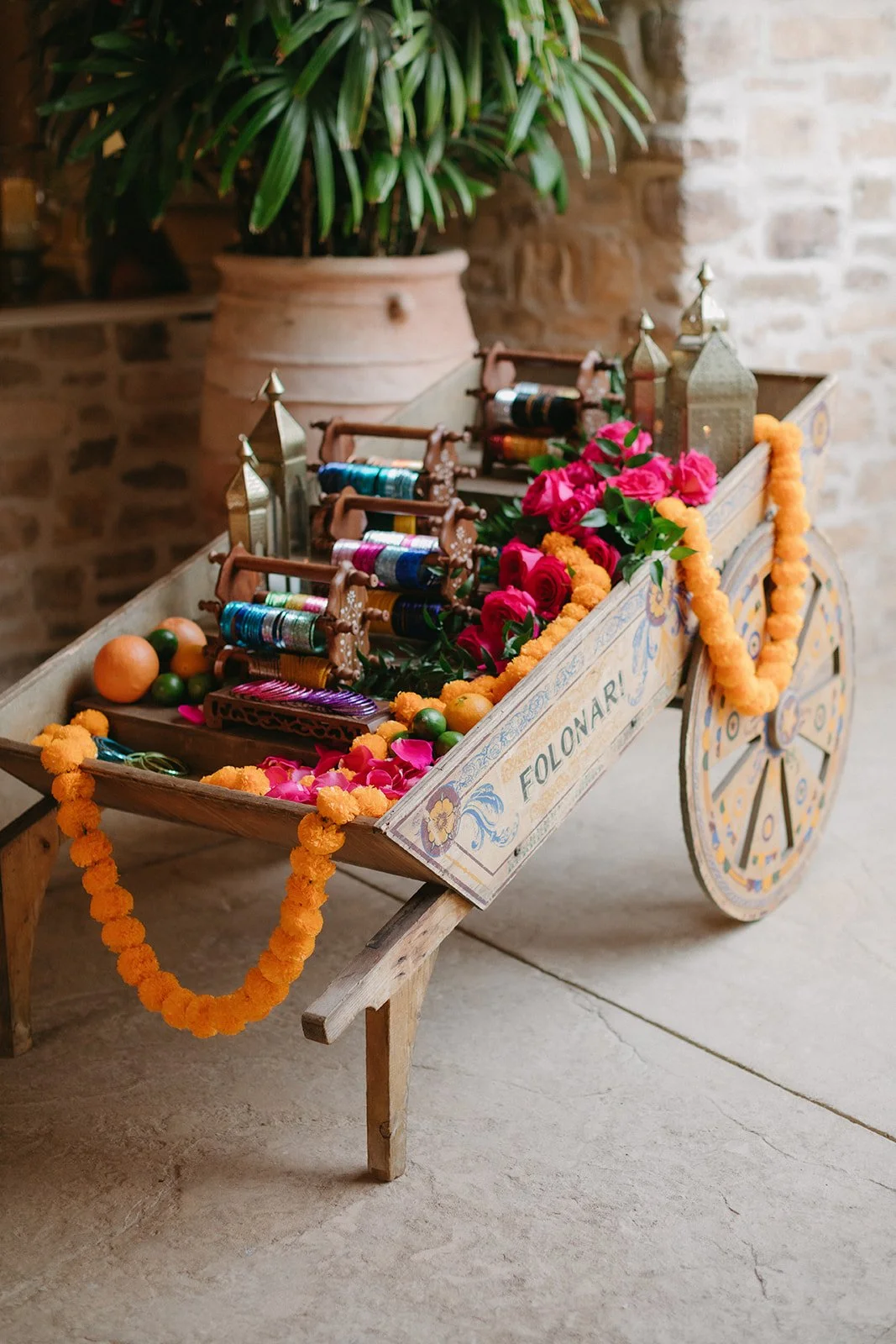 A decorative wooden cart filled with colorful flowers, marigold garlands, and spools of thread, with a potted plant in the background.
