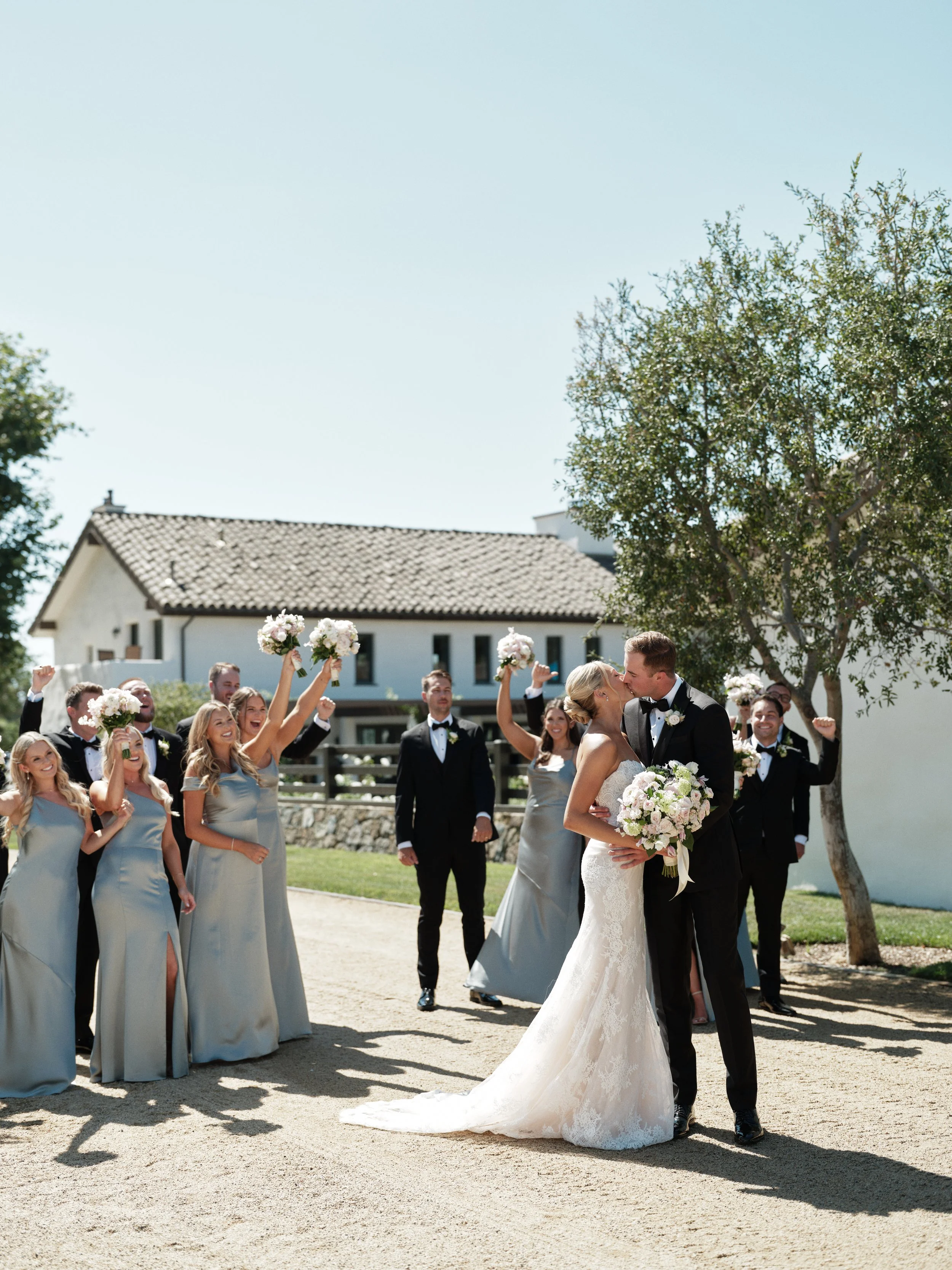 Bride and groom kissing while wedding party celebrates around them outside on a sunny day.