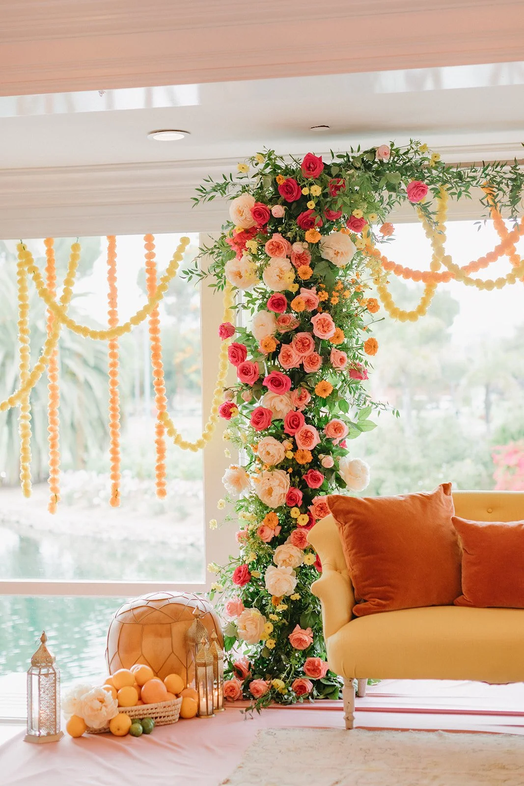 Decorative floral arrangement with pink, white, and red roses and yellow flowers on a vertical stand, with peach-colored pillows on a cream-colored sofa and yellow and orange garlands hanging in the background.