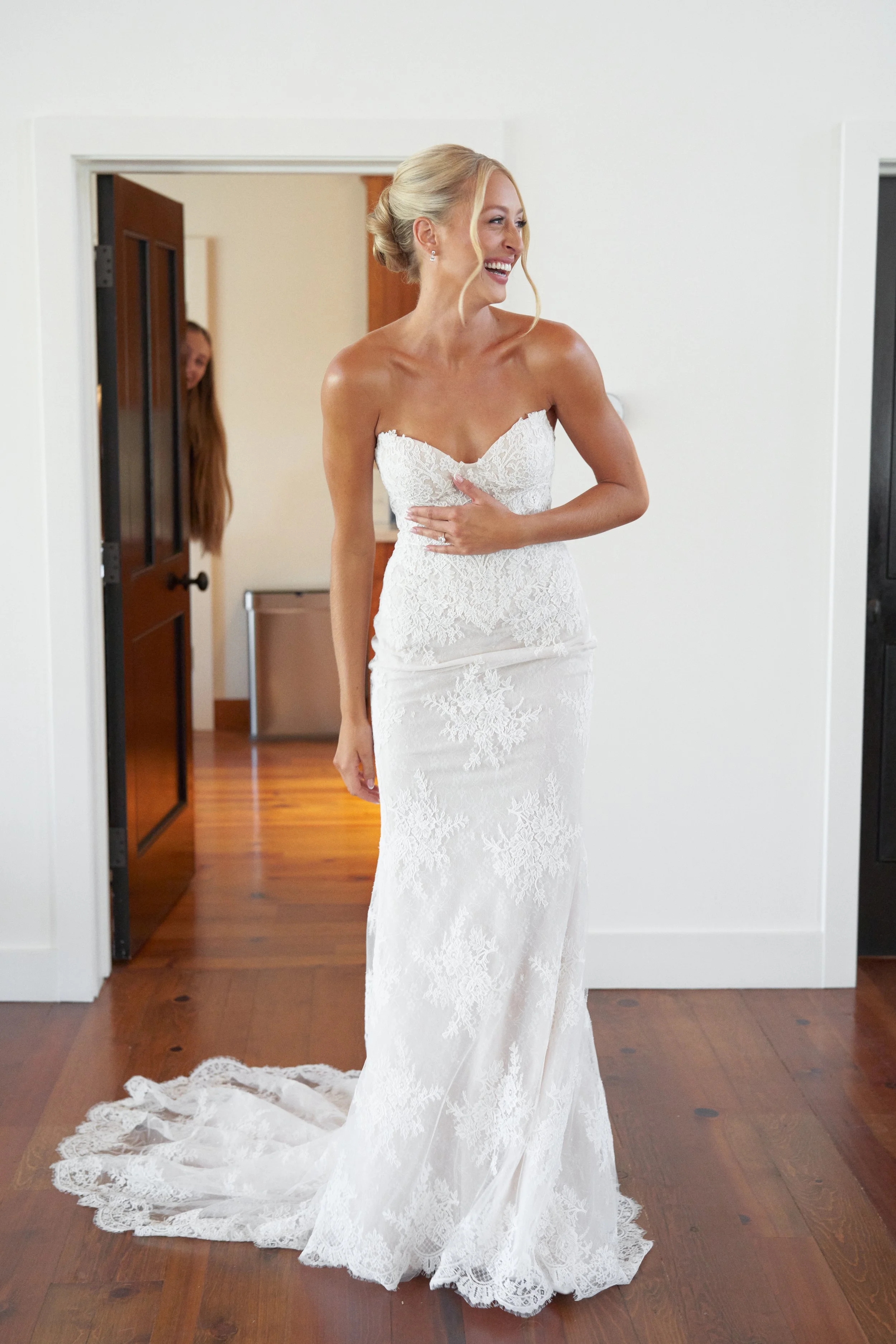 A bride in a white lace wedding gown standing inside, smiling as she looks to the side, with a woman peeking through an open door in the background.