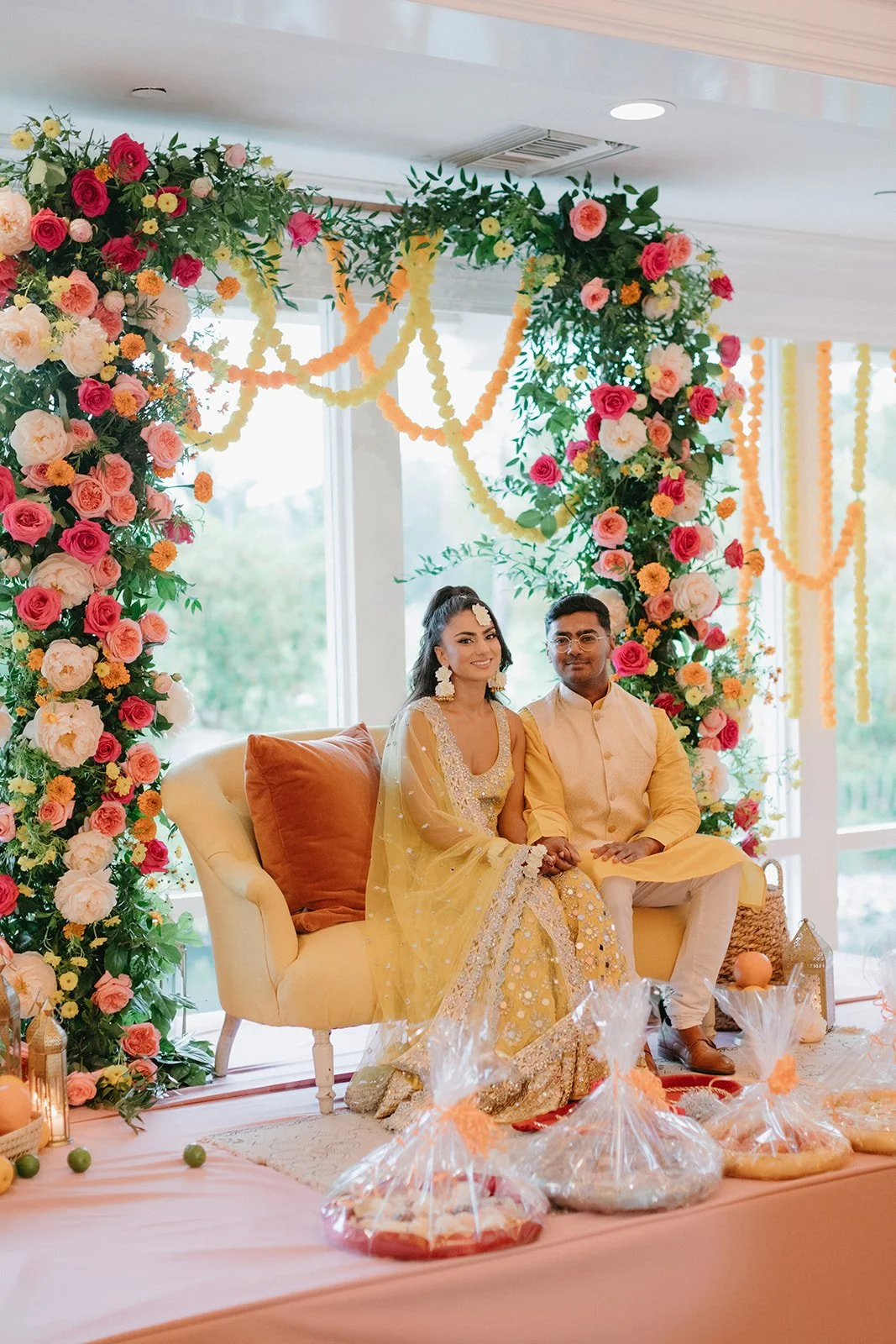 A newlywed Indian couple sits on a yellow sofa, holding hands, surrounded by floral decorations and wedding items at their celebration.