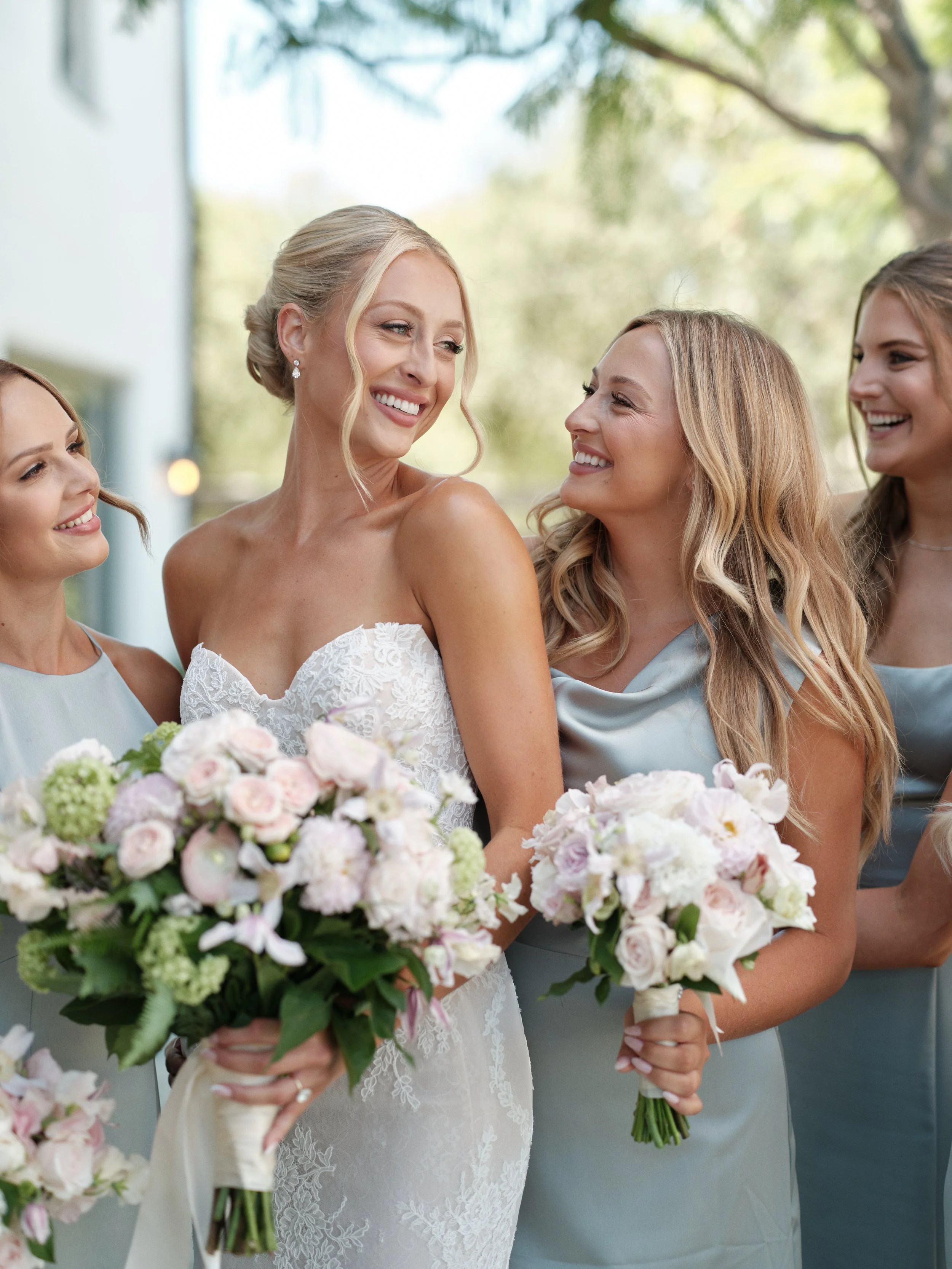Bride with blonde hair in wedding dress holding a bouquet, smiling with bridesmaids.