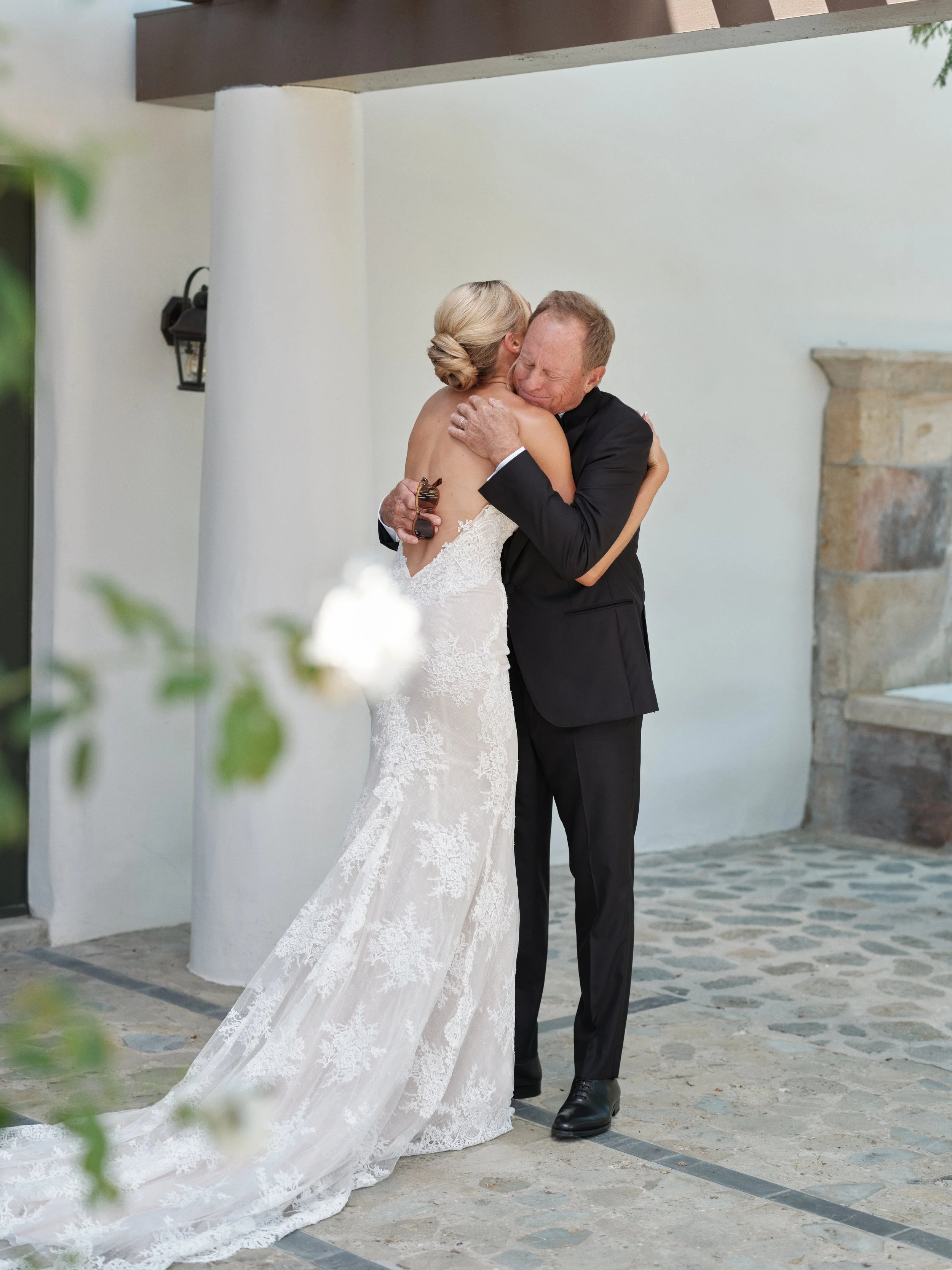 A bride in a lace wedding gown embraces an older man in a black suit, who is also hugging her tightly, outside a building with white walls and stone accents.
