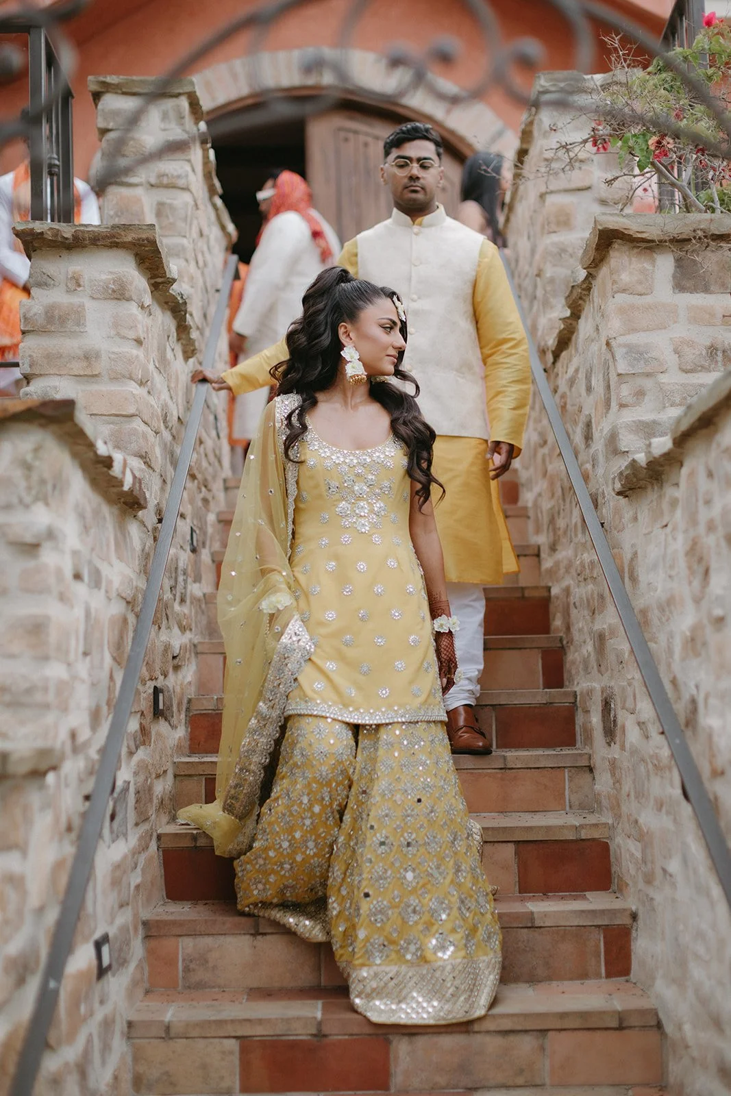 A woman dressed in a yellow embroidered traditional Indian outfit is descending a stone staircase. A man behind her is wearing a cream and yellow traditional outfit. The setting appears to be a rustic, outdoor location with stone walls and wooden acc
