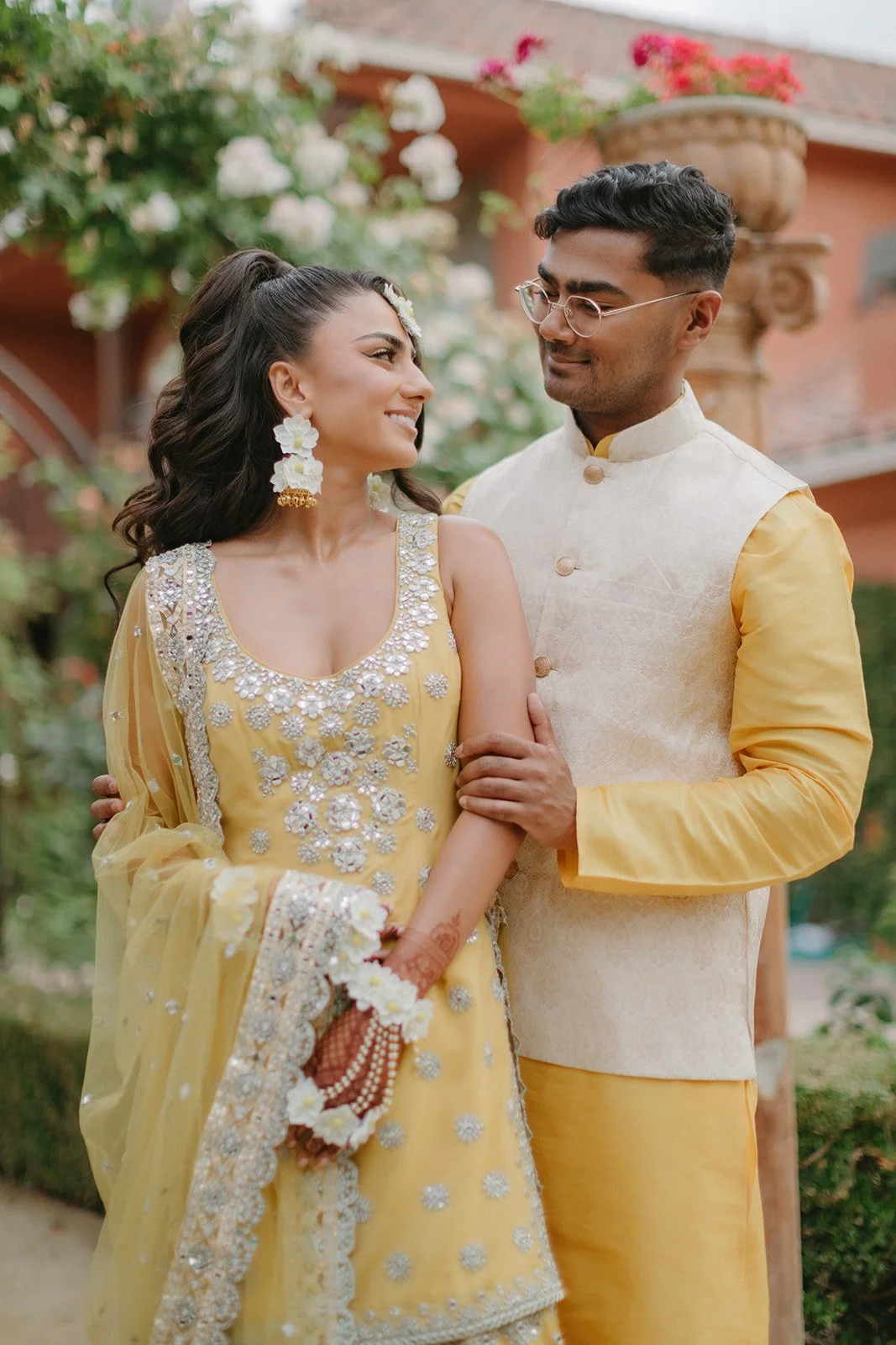 A couple dressed in traditional Indian attire standing outdoors, with the woman gazing at the man while he smiles at her. The woman wears a yellow embroidered outfit with floral jewelry, and the man wears a cream-colored vest with yellow sleeves.