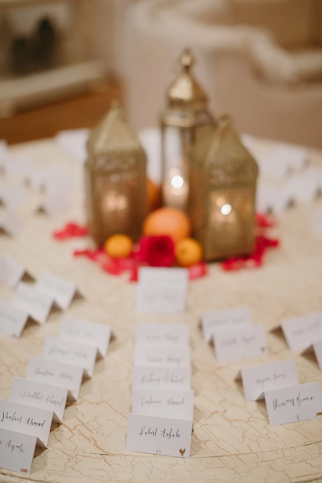 Wedding seating chart with handwritten names on white cards, decorated with lanterns, candles, and fruits in the background.