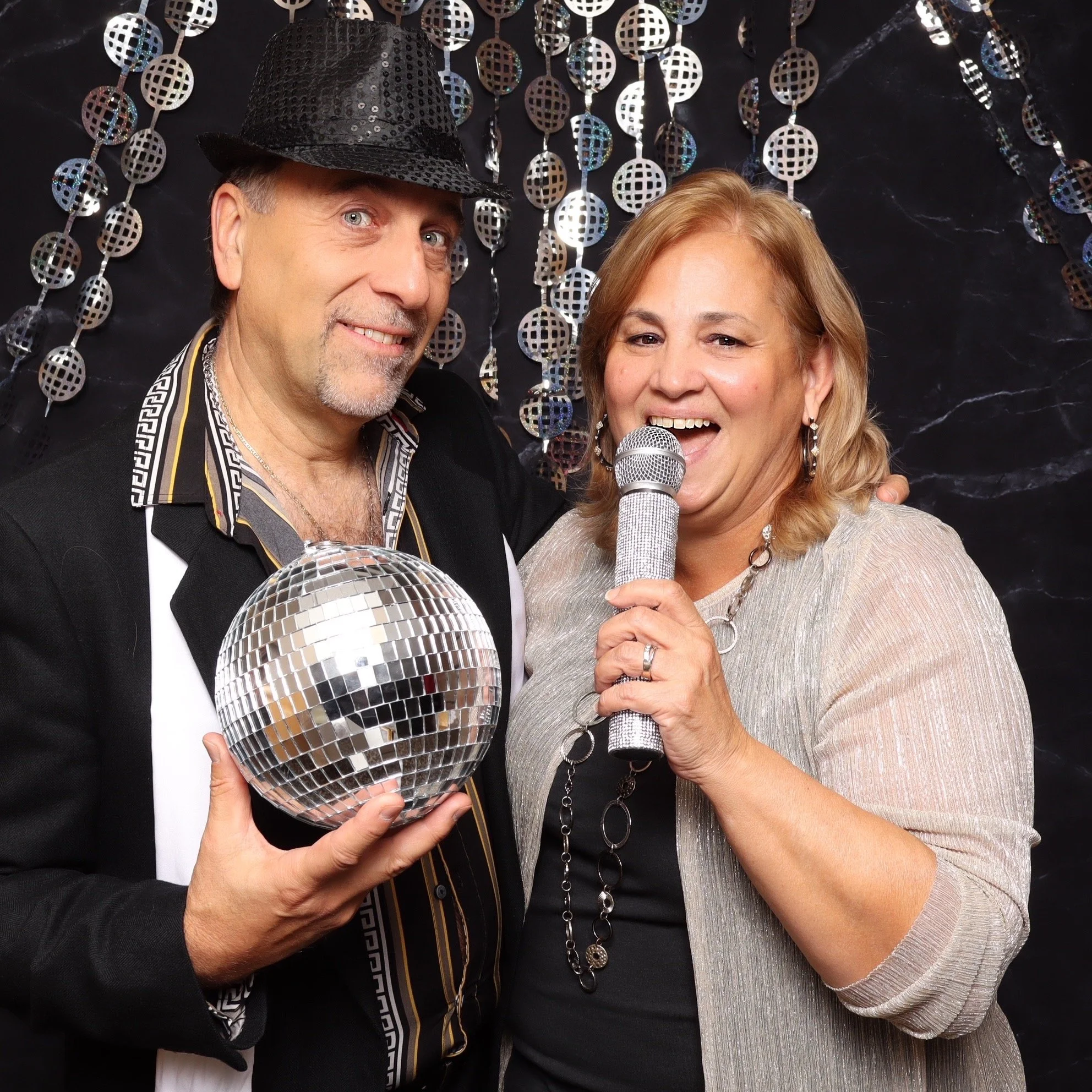 A man in a black shirt and hat holding a disco ball, standing next to a smiling woman holding a microphone during a celebration.