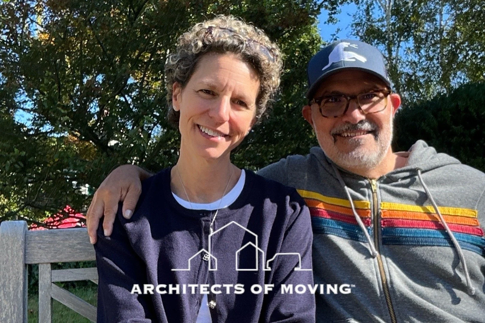 A smiling woman with short curly hair and a man with glasses and a cap sitting together outside on a sunny day, with trees in the background. The woman is wearing a dark jacket and a necklace, and the man is wearing a gray hoodie with colorful stripe