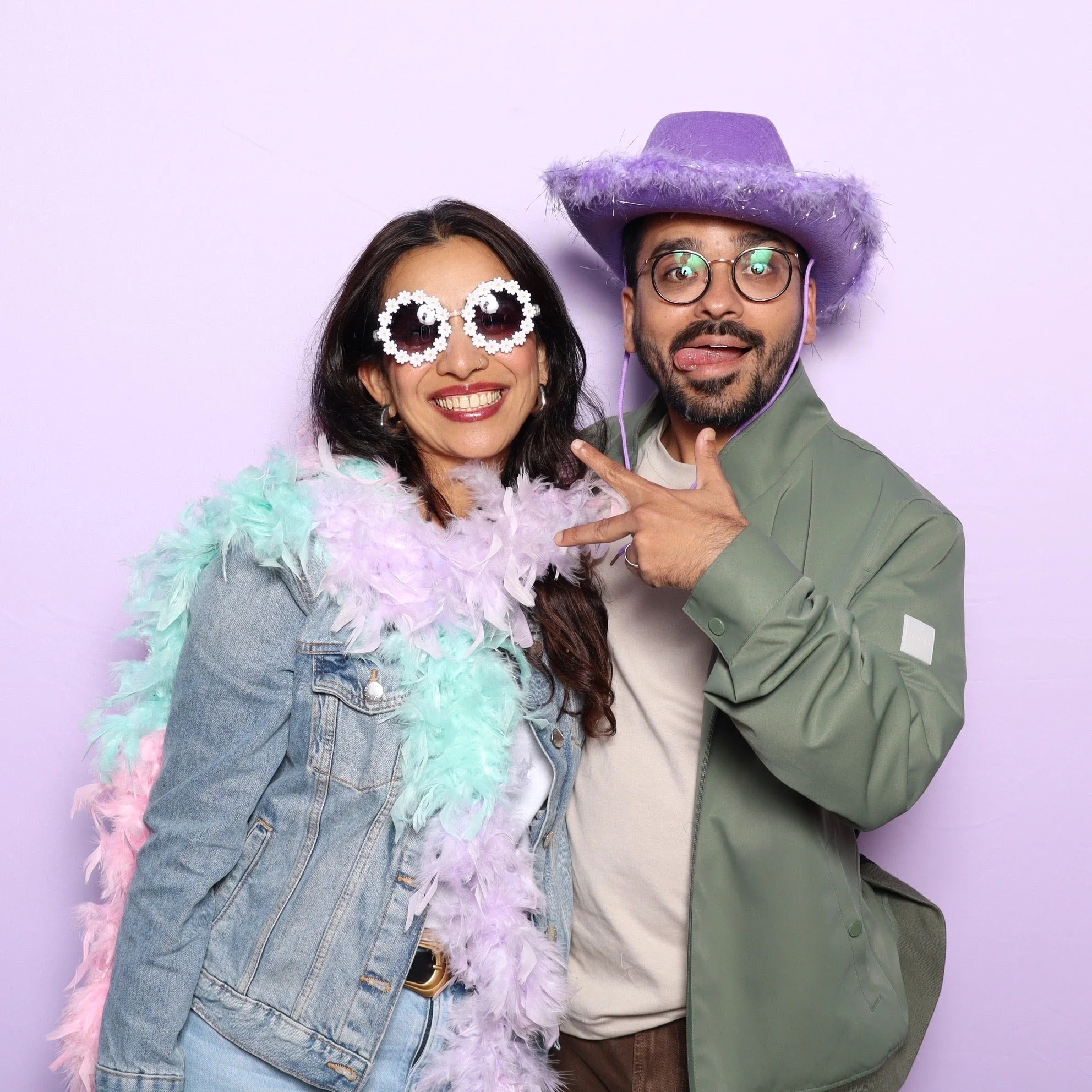 Two people in colorful costumes pose playfully against a light purple background. The woman on the left wears feathered accessories, denim jacket, and sunglasses, while the man on the right wears a purple cowboy hat, glasses, and a peace sign gesture