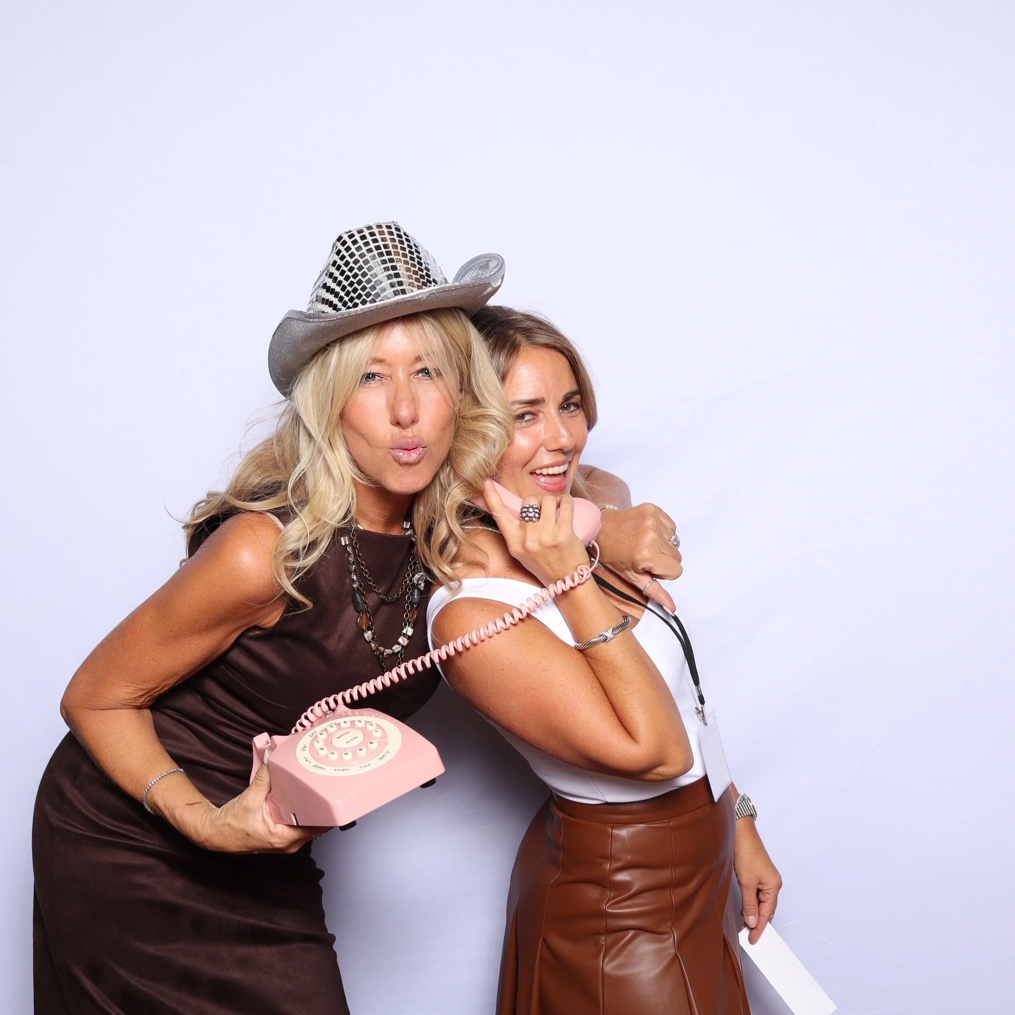 Two women posing playfully, one wearing a silver hat and holding a pink rotary phone, both smiling and making kiss faces against a plain white background.