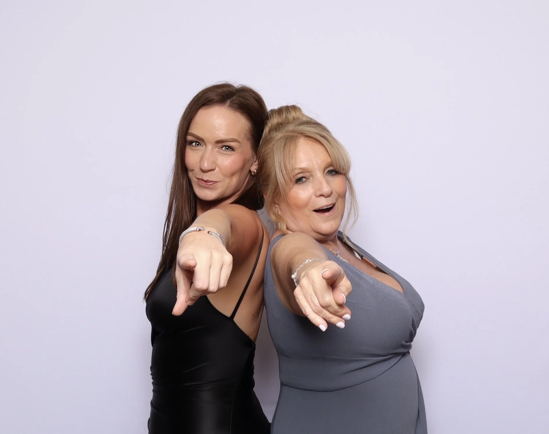Two women with blonde and brunette hair pointing at the camera, smiling, against a plain white background.