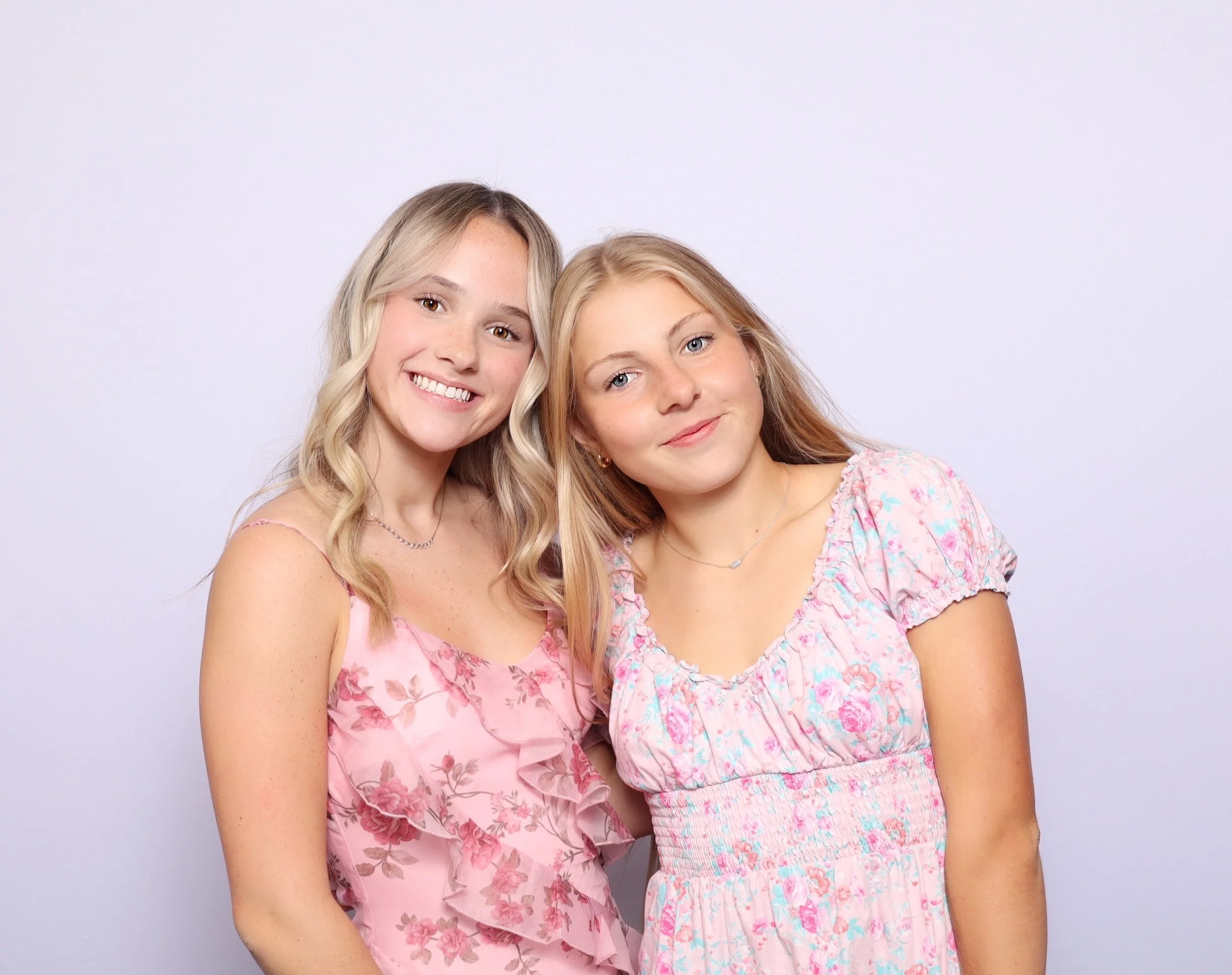 Two young women with blonde hair smiling and standing close together against a plain white background, both wearing pink floral dresses.