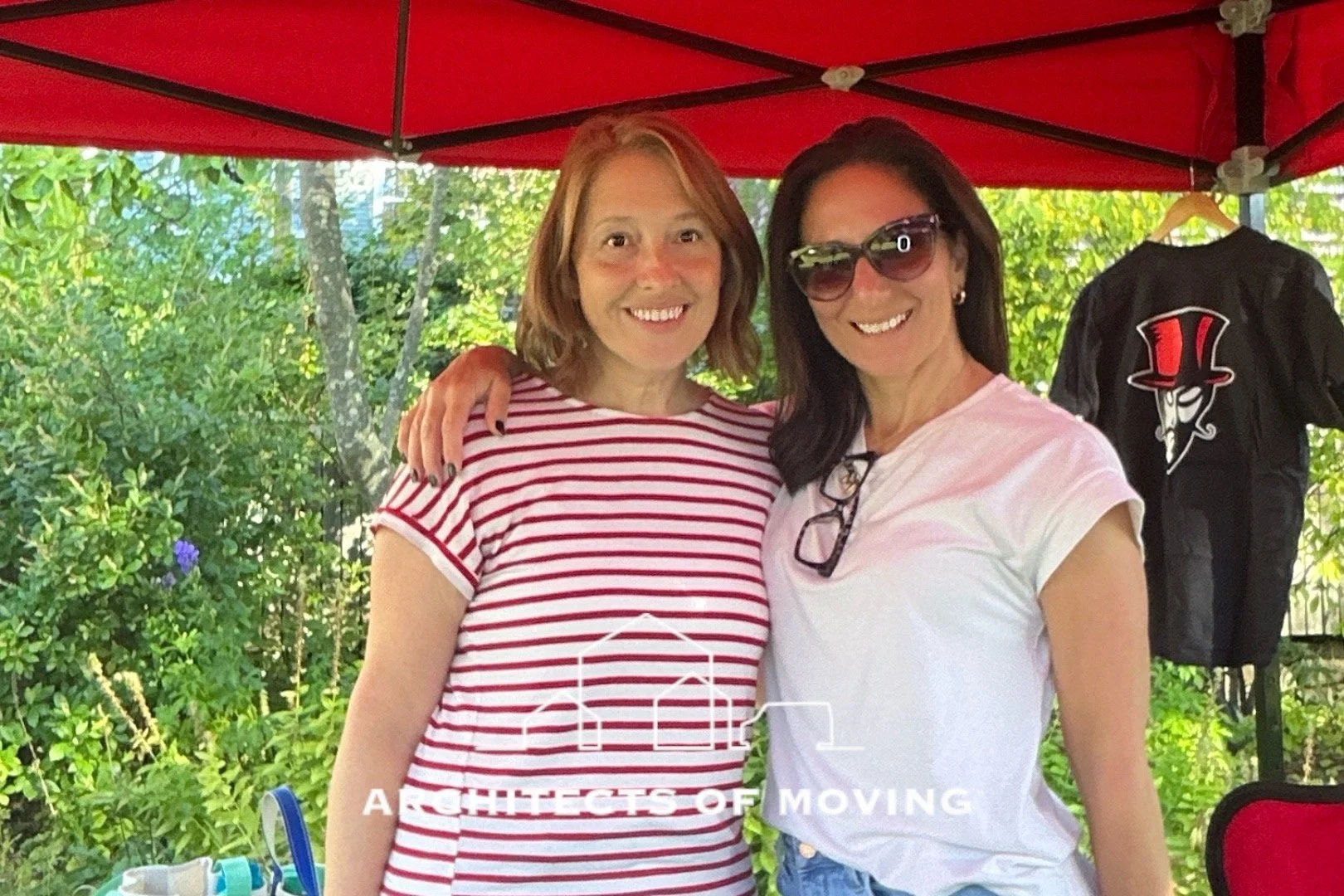 Two women smiling, one in a red and white striped shirt and the other in a white shirt, standing close under a red canopy, with greenery outside and a black jacket with a graphic of a top hat and face on a hanger in the background.