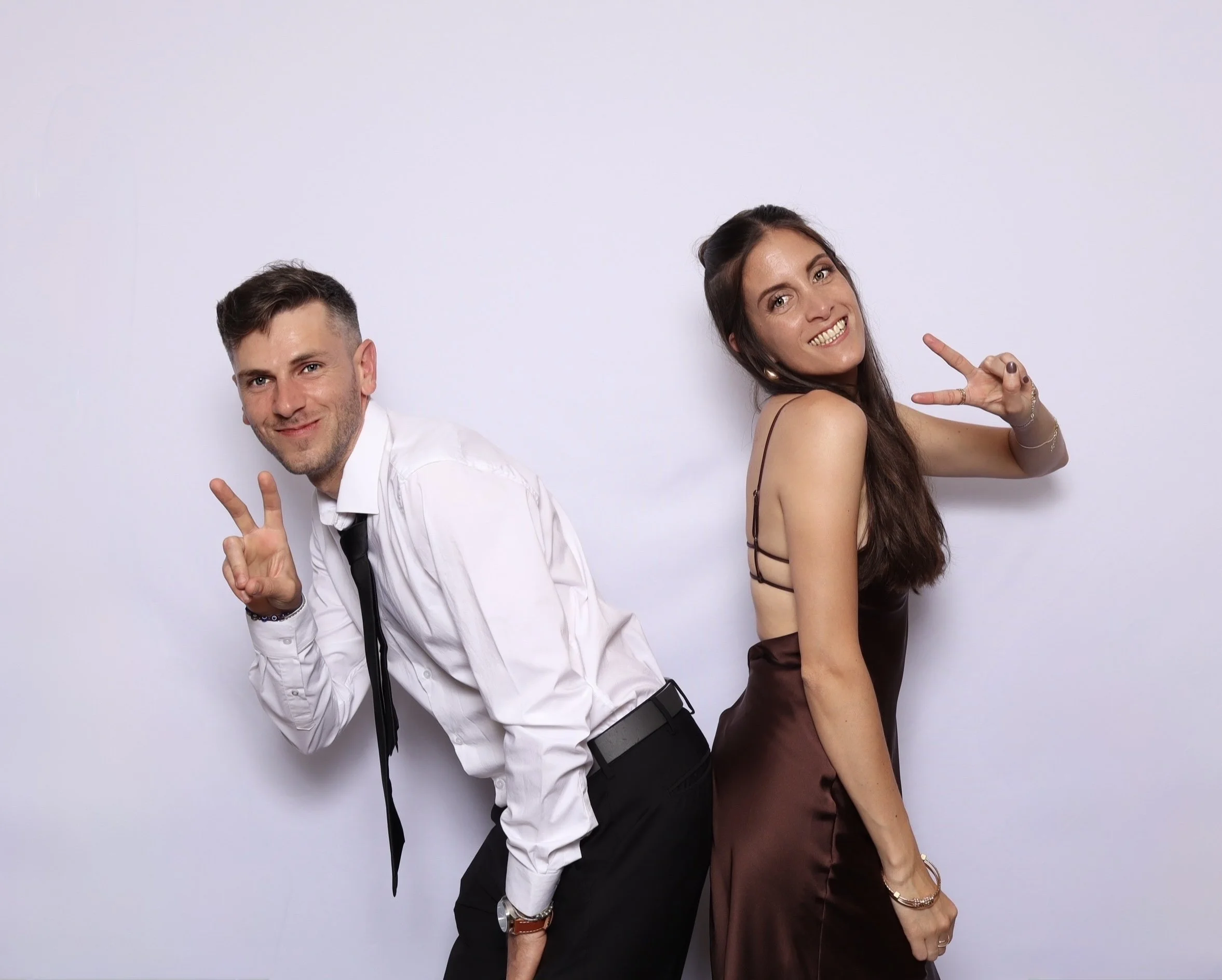 A man and woman standing back to back against a plain white wall, smiling and making peace signs with their hands.