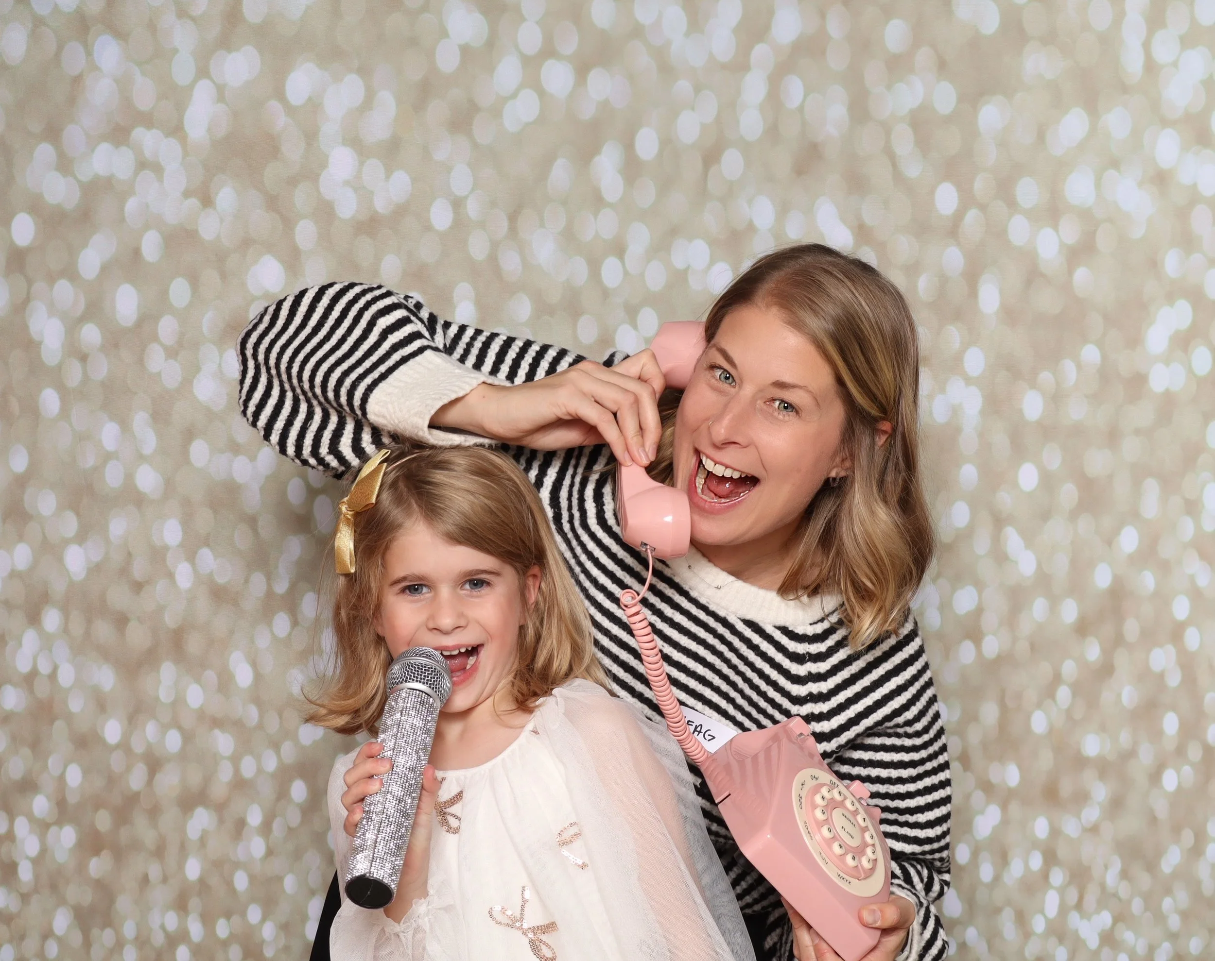 A woman and a young girl are posing playfully, with the woman holding a pink vintage rotary phone to her ear and the girl holding a microphone. They are smiling and appear to be having fun against a bokeh background.