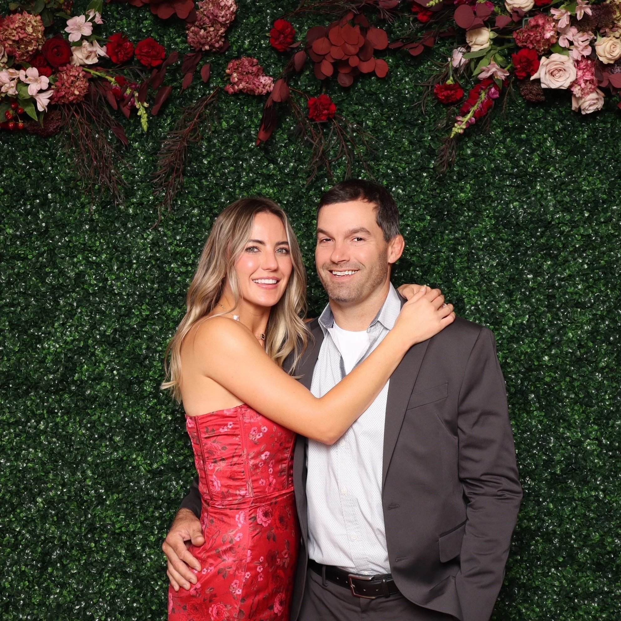 A smiling couple poses together in front of a decorative green leaf wall with pink and red flowers at the top. The woman has long blonde hair, wears a red floral strapless dress, and has her arm around the man's shoulder. The man has short dark hair,