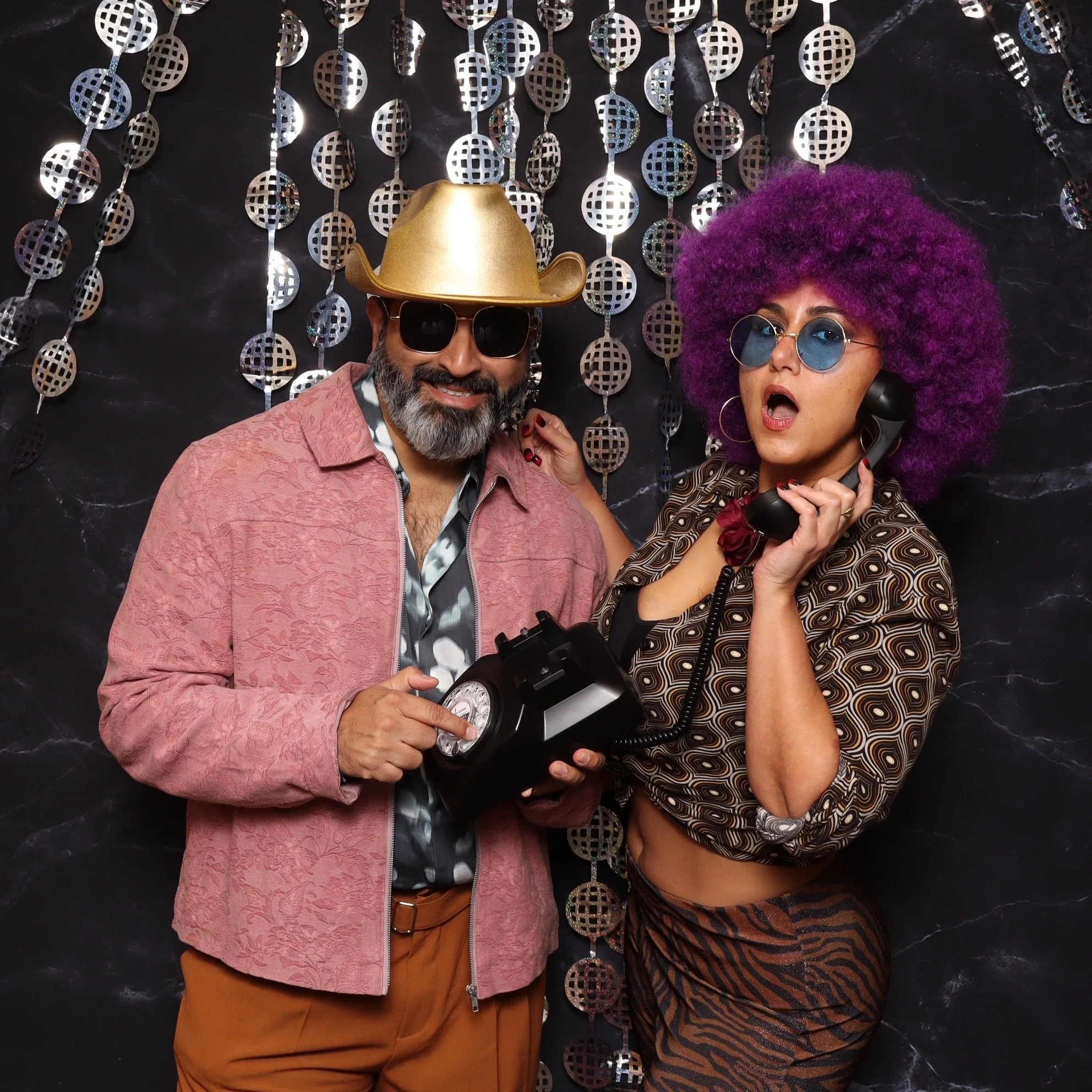 Two people in retro outfits with colorful accessories posing in front of a shiny, metallic backdrop.
