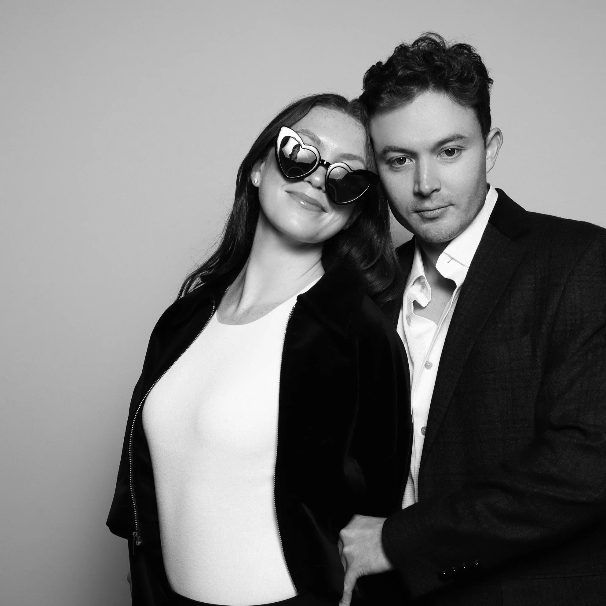 A black-and-white photo of a smiling woman wearing sunglasses and a dark jacket, standing close to a man in a suit, both posing against a plain background.