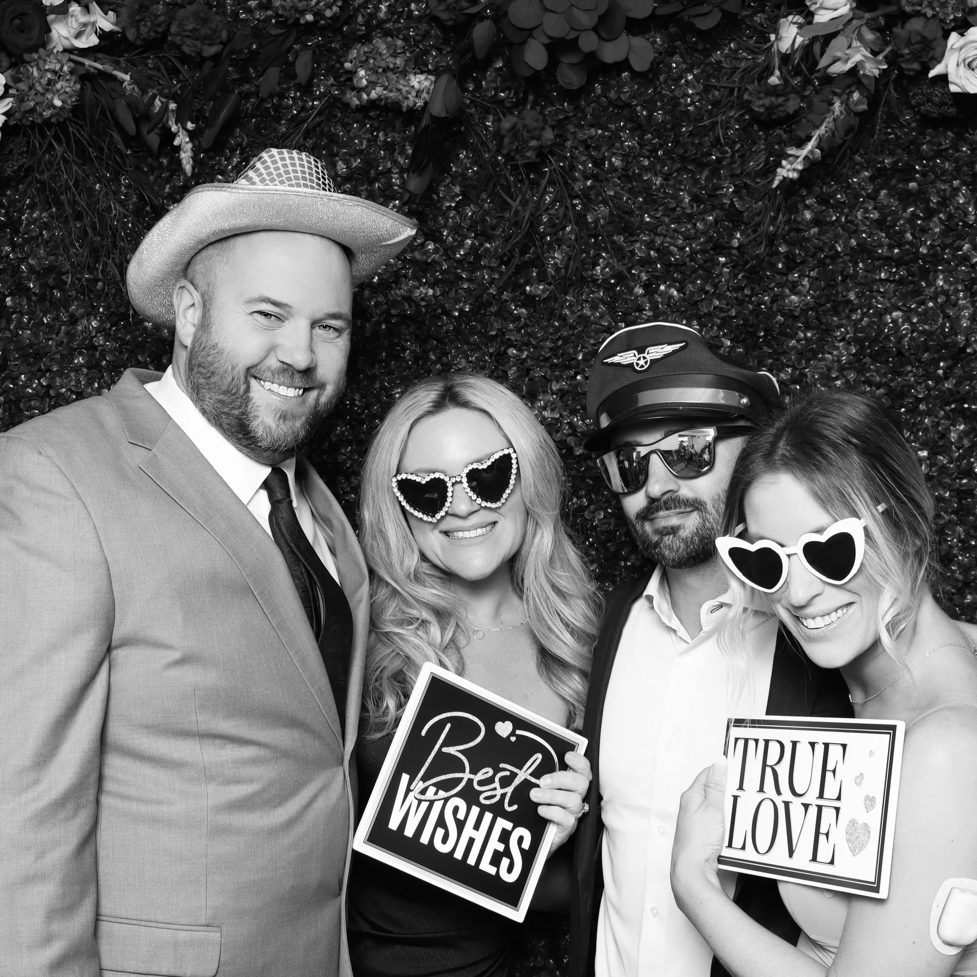 Four people smiling in a photo booth wearing fun accessories, holding signs that say "Best Wishes" and "True Love," against a leafy backdrop.