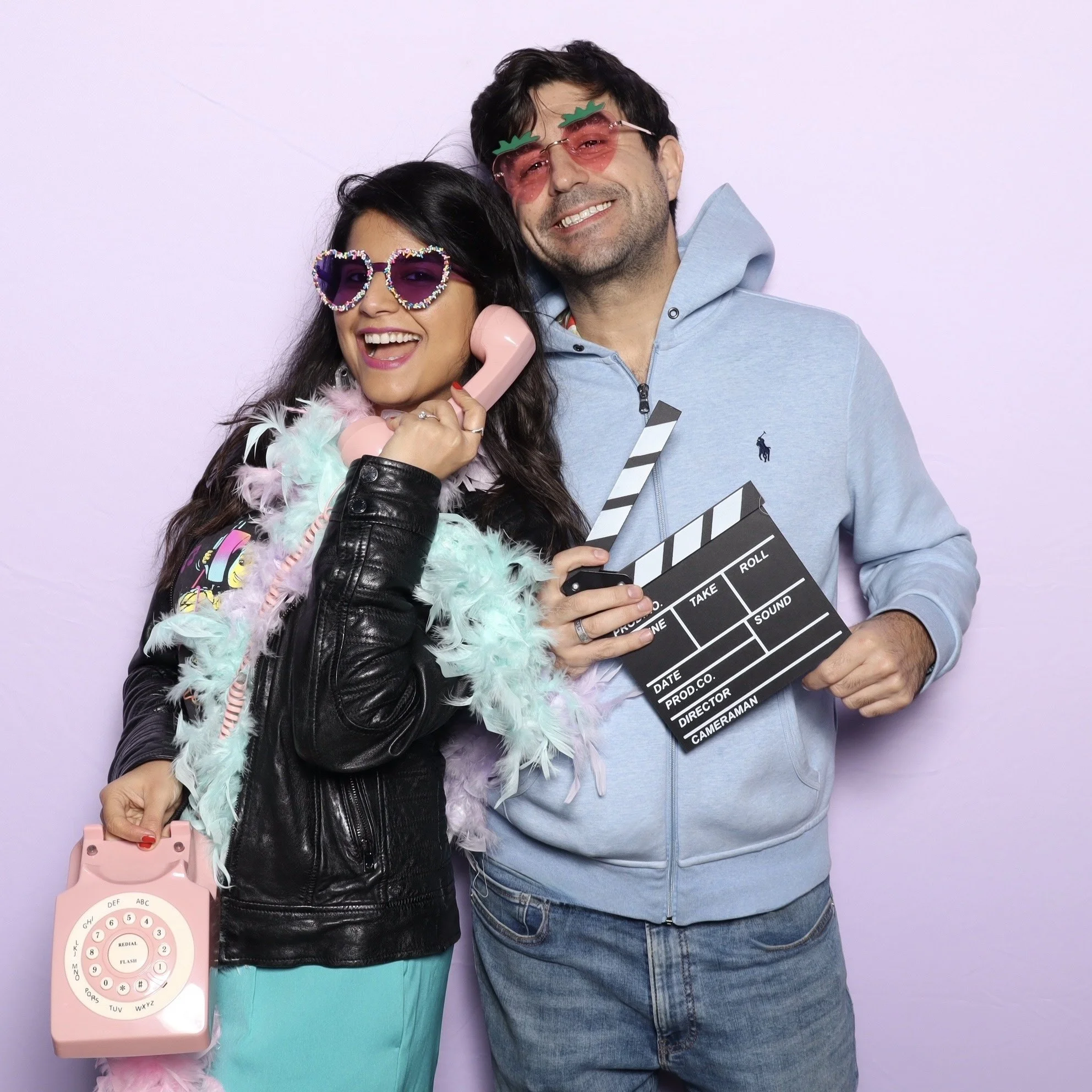 Two people dressed in colorful, playful costumes holding filming props are smiling and posing in front of a light purple background.