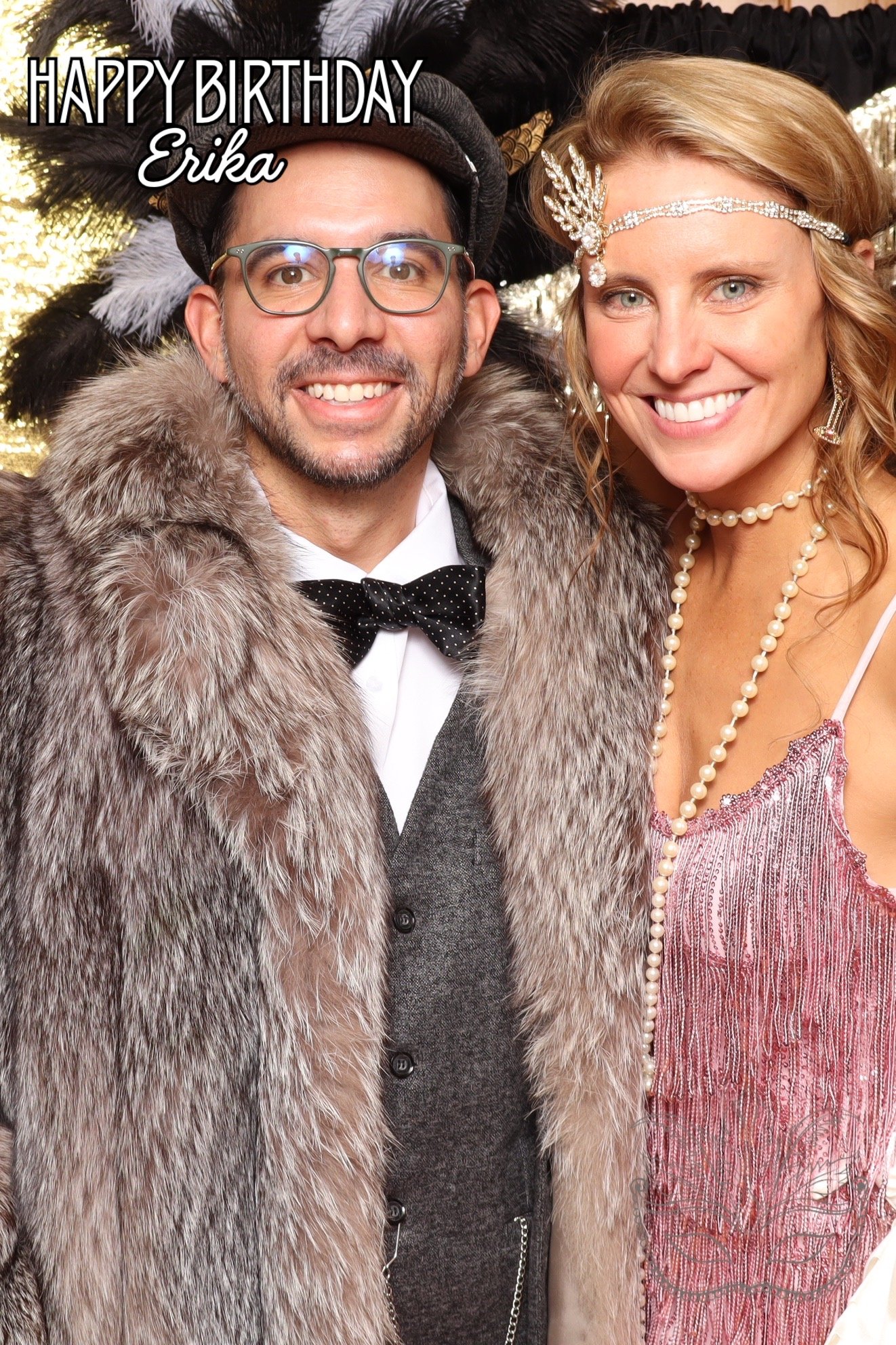 A man in a tuxedo with a fur coat and hat, and a woman in a pink dress with pearls, smiling at a birthday celebration with a backdrop of a decorated Christmas tree. The image has the text 'HAPPY BIRTHDAY Erika'.