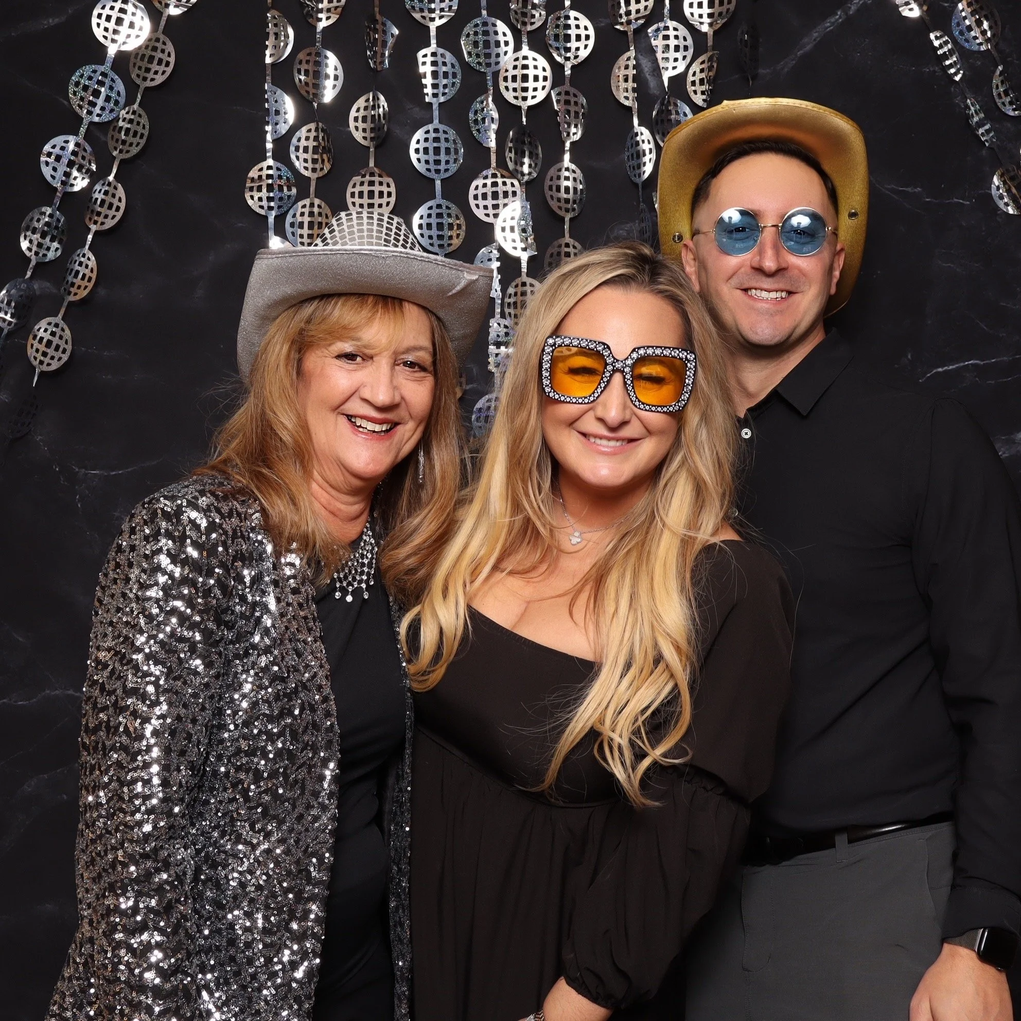 Three people dressed in festive outfits, wearing sunglasses and hats, smiling at a party with a metallic disco ball background.