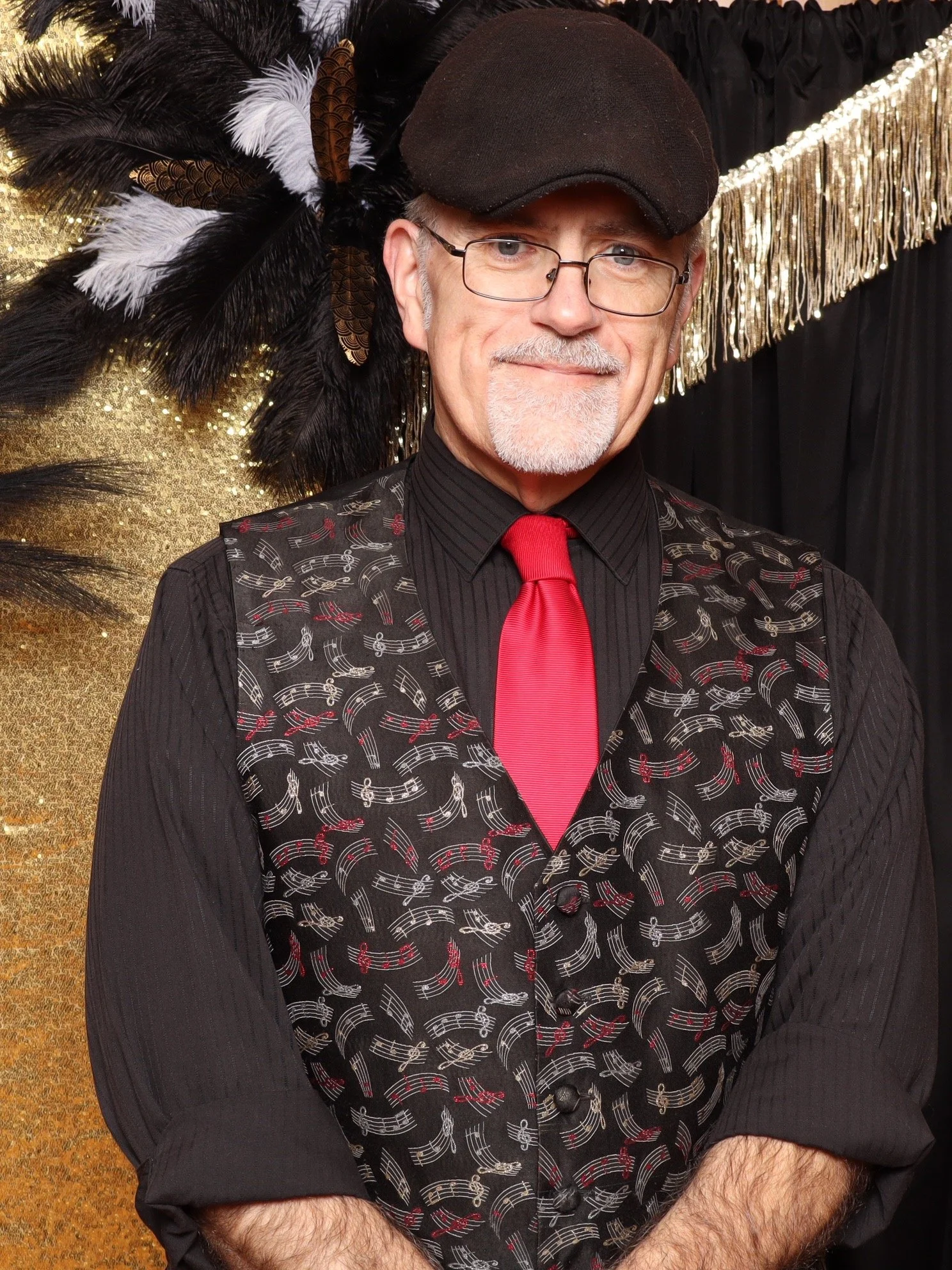 A man wearing glasses, a black hat, a black shirt with a red tie, and a black vest with musical notes, standing in front of a black and gold backdrop with feathers and fringe.