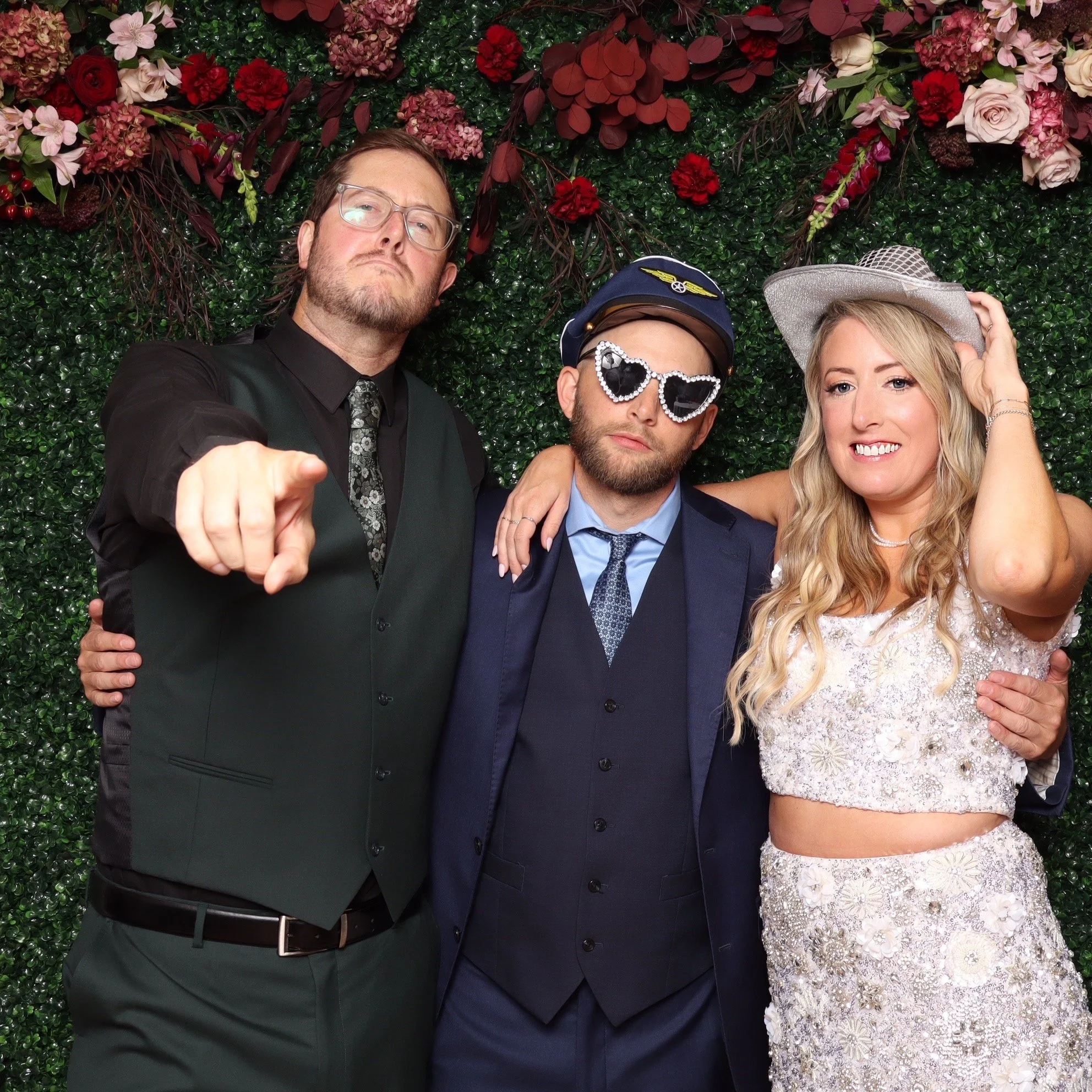 Three people posing for a photo in front of a floral green backdrop; a man on the left pointing and wearing glasses, a dark suit with a vest, and a necktie; in the middle, a man with a beard wearing a police officer hat, heart-shaped sunglasses, and 