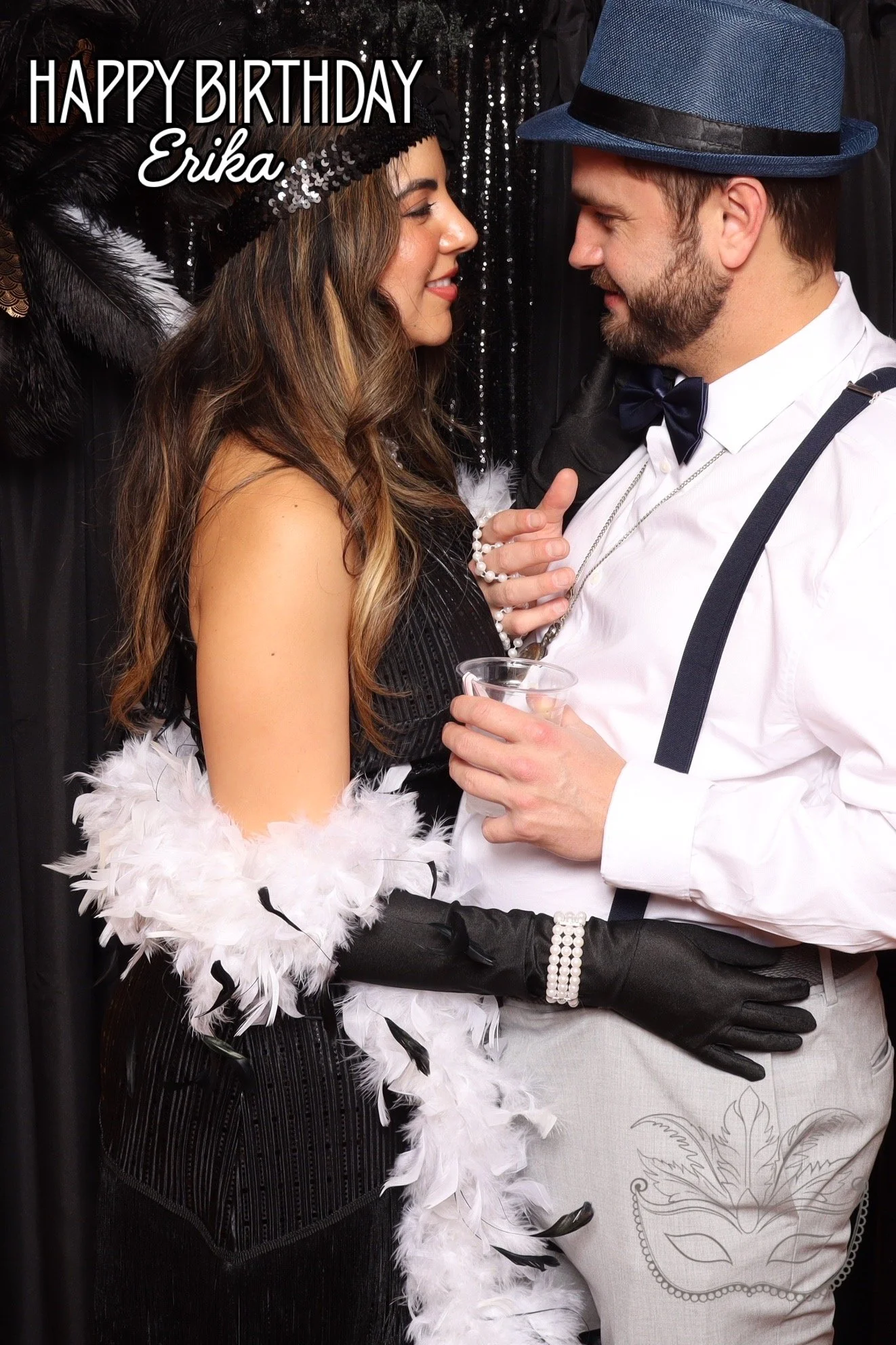 A couple dressed in vintage-style clothing at a party, with the woman wearing a feathered hat and boa and the man wearing a hat, bow tie, and suspenders, holding a drink, in front of a black background with the text "Happy Birthday Erika" in the top 